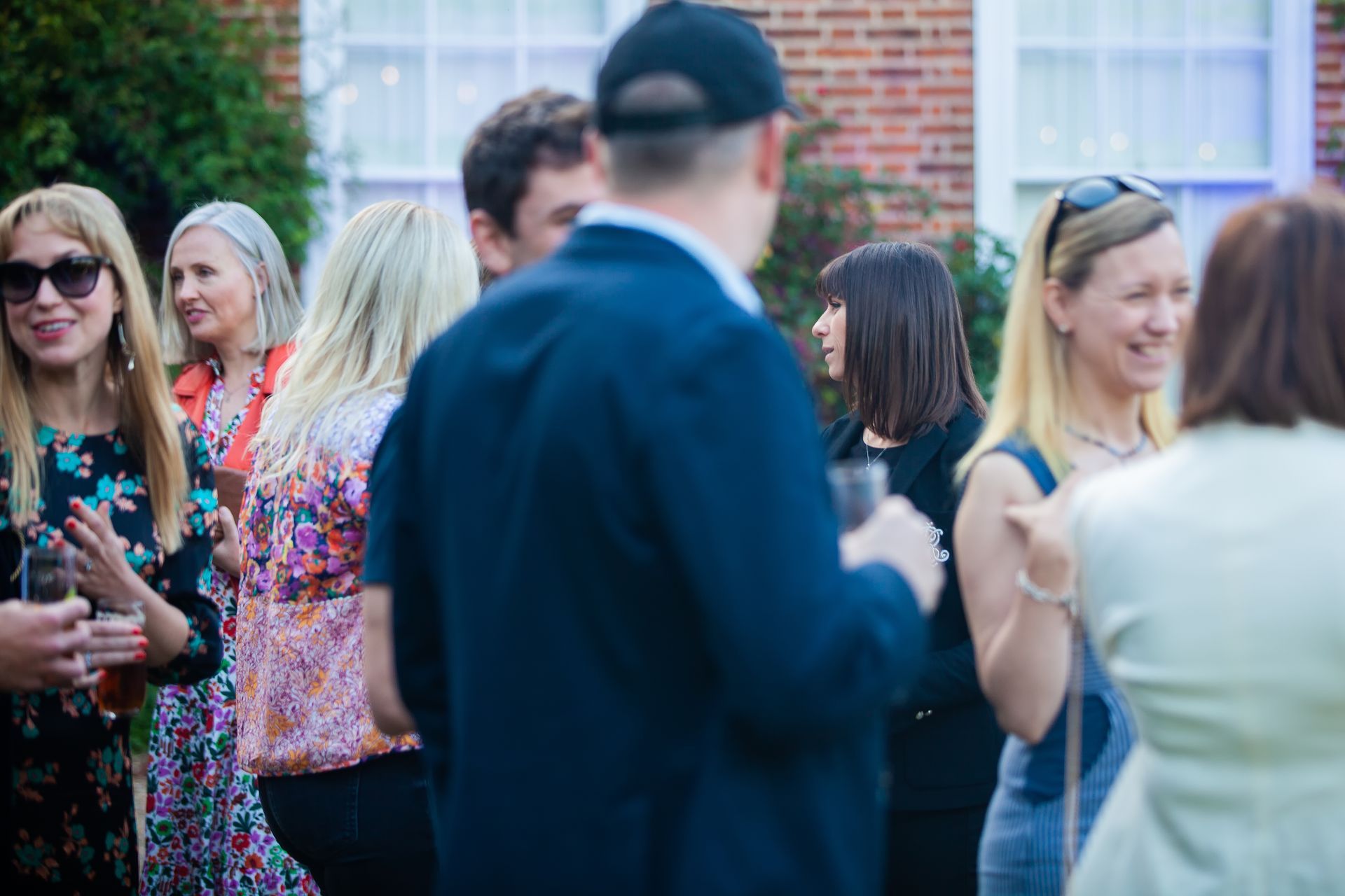 People socializing outdoors, some holding drinks, near a brick building.
