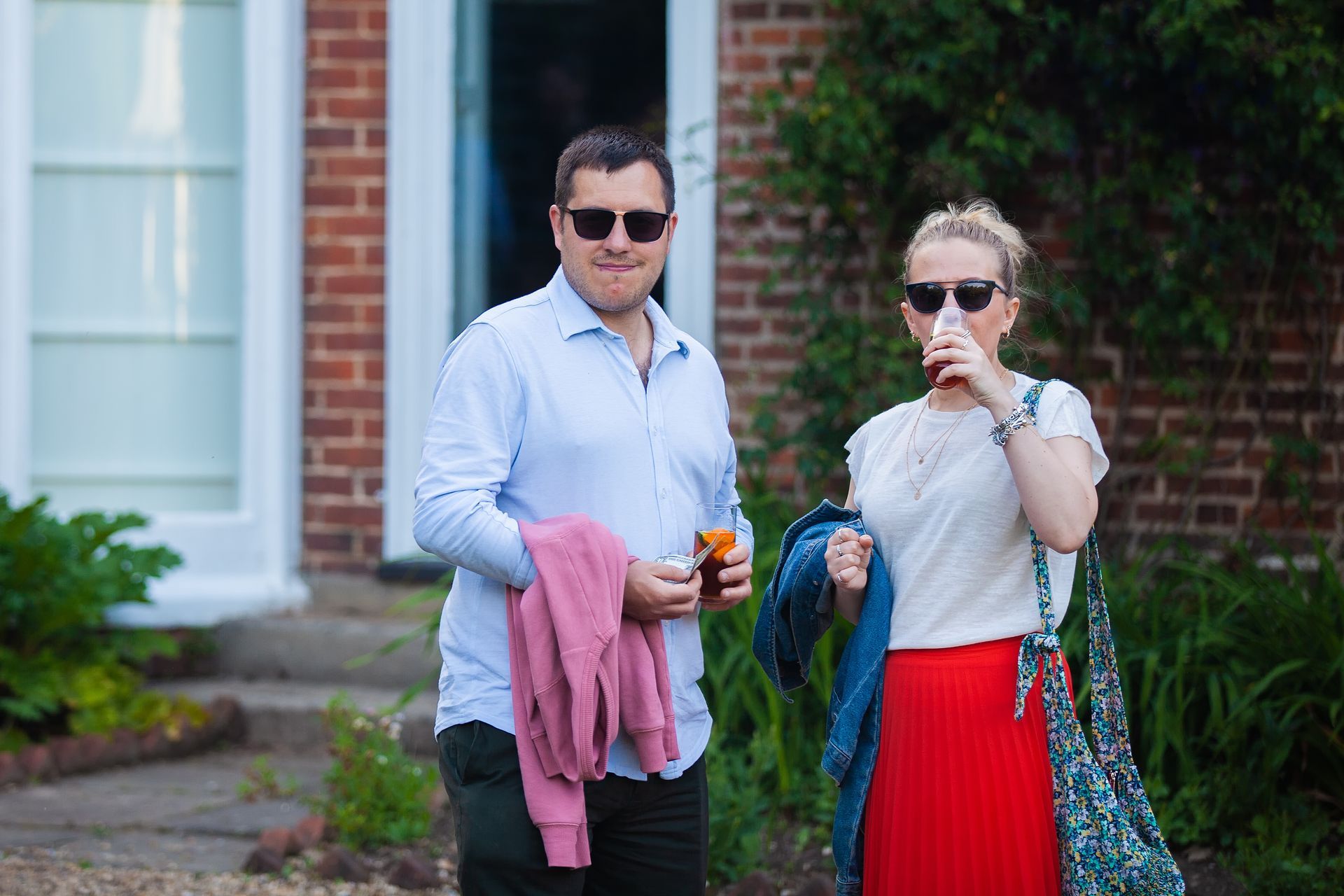 Man and woman wearing sunglasses, standing outside a brick building. The woman is holding a drink, the man a sweater.