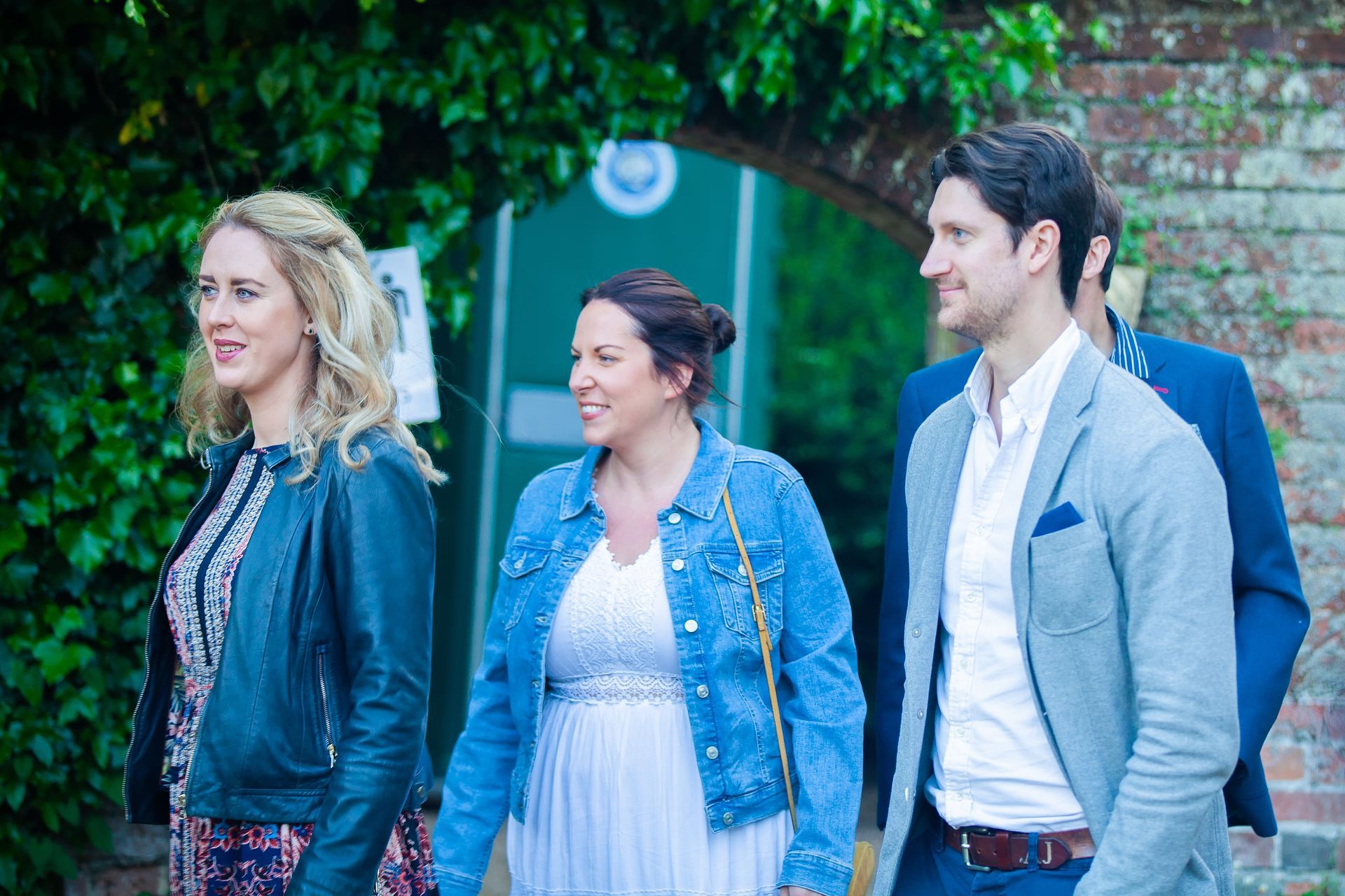 Four people walking towards a brick archway with a tennis court sign; smiling, outdoors.