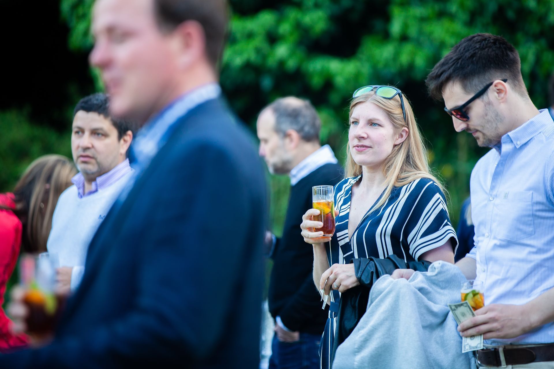 People at an outdoor event, some holding drinks. A woman in a striped top looks toward the right.