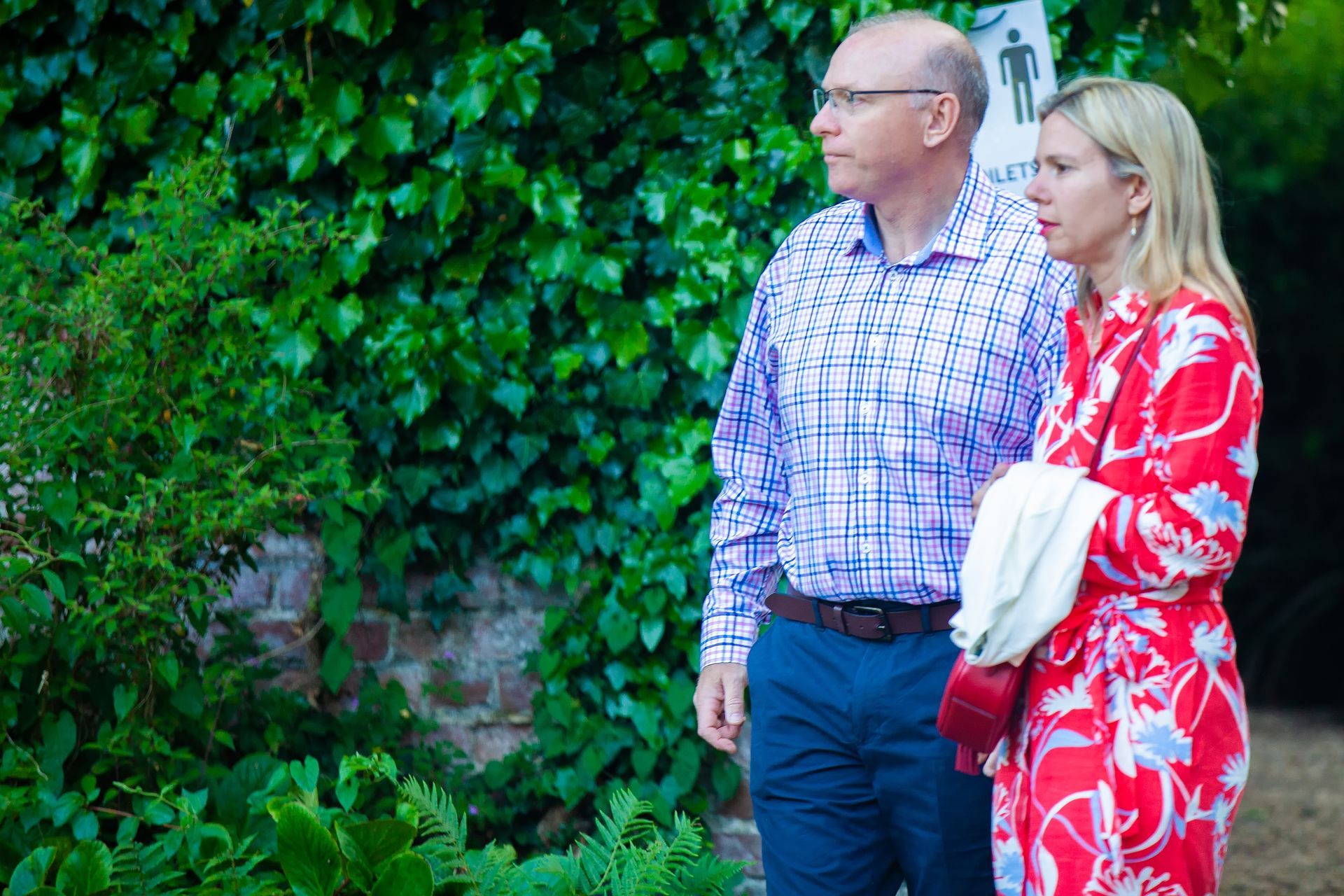 Man and woman looking to the side; man in checkered shirt, woman in red floral dress, in a garden setting.
