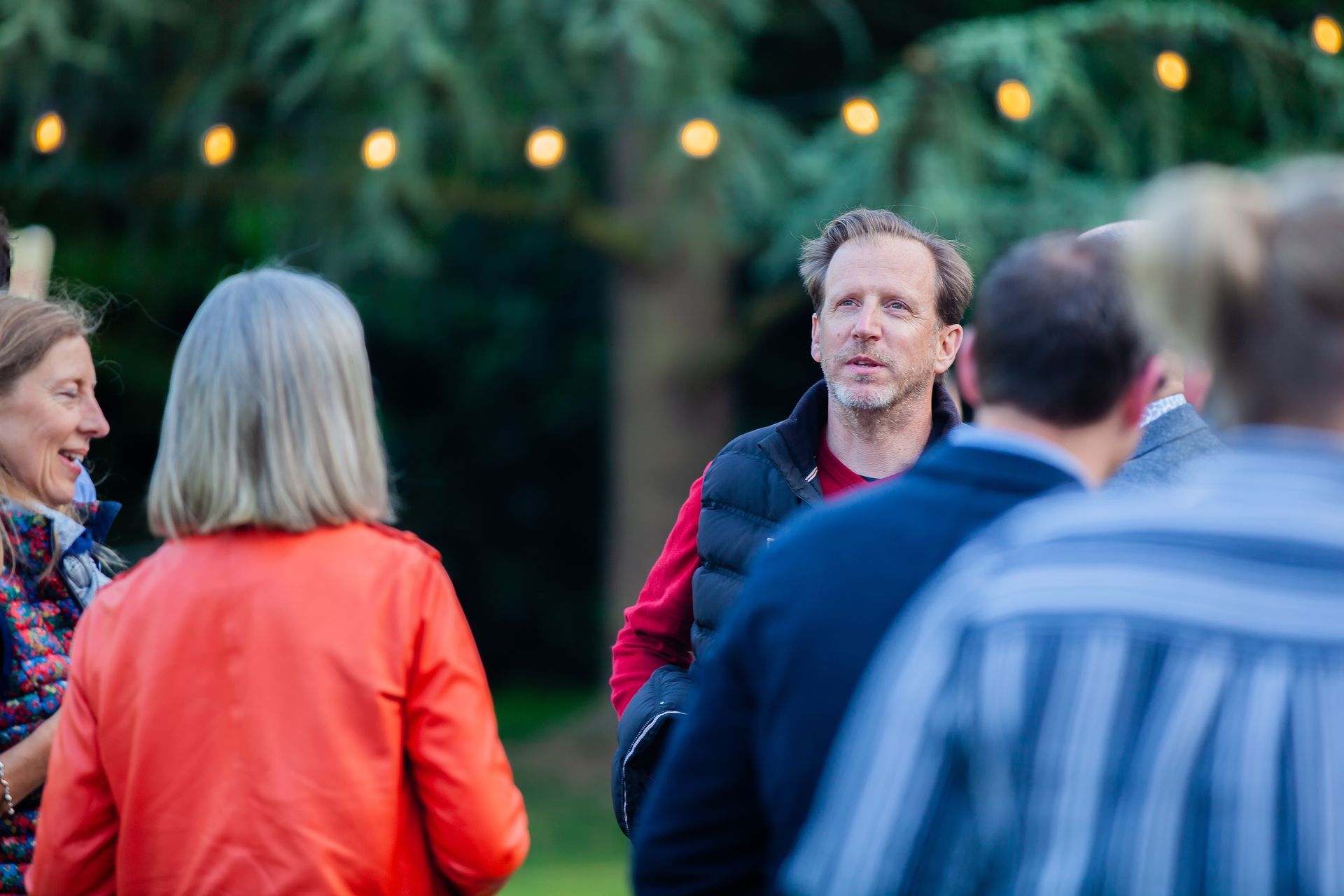 People socializing outdoors; man in red shirt looks up, others in background, string lights.