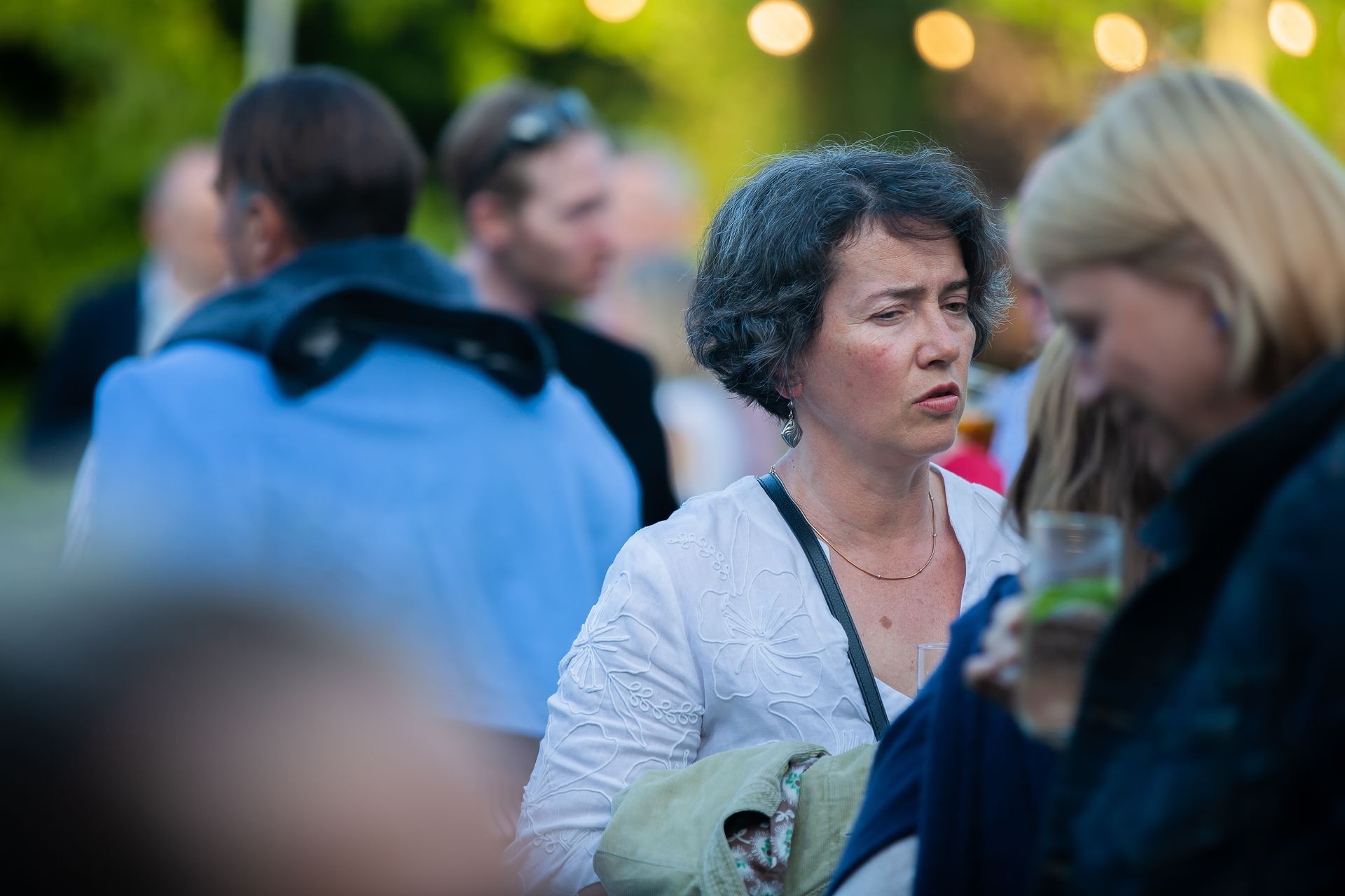 Woman with curly gray hair at an outdoor gathering, looking concerned, wearing a white top.
