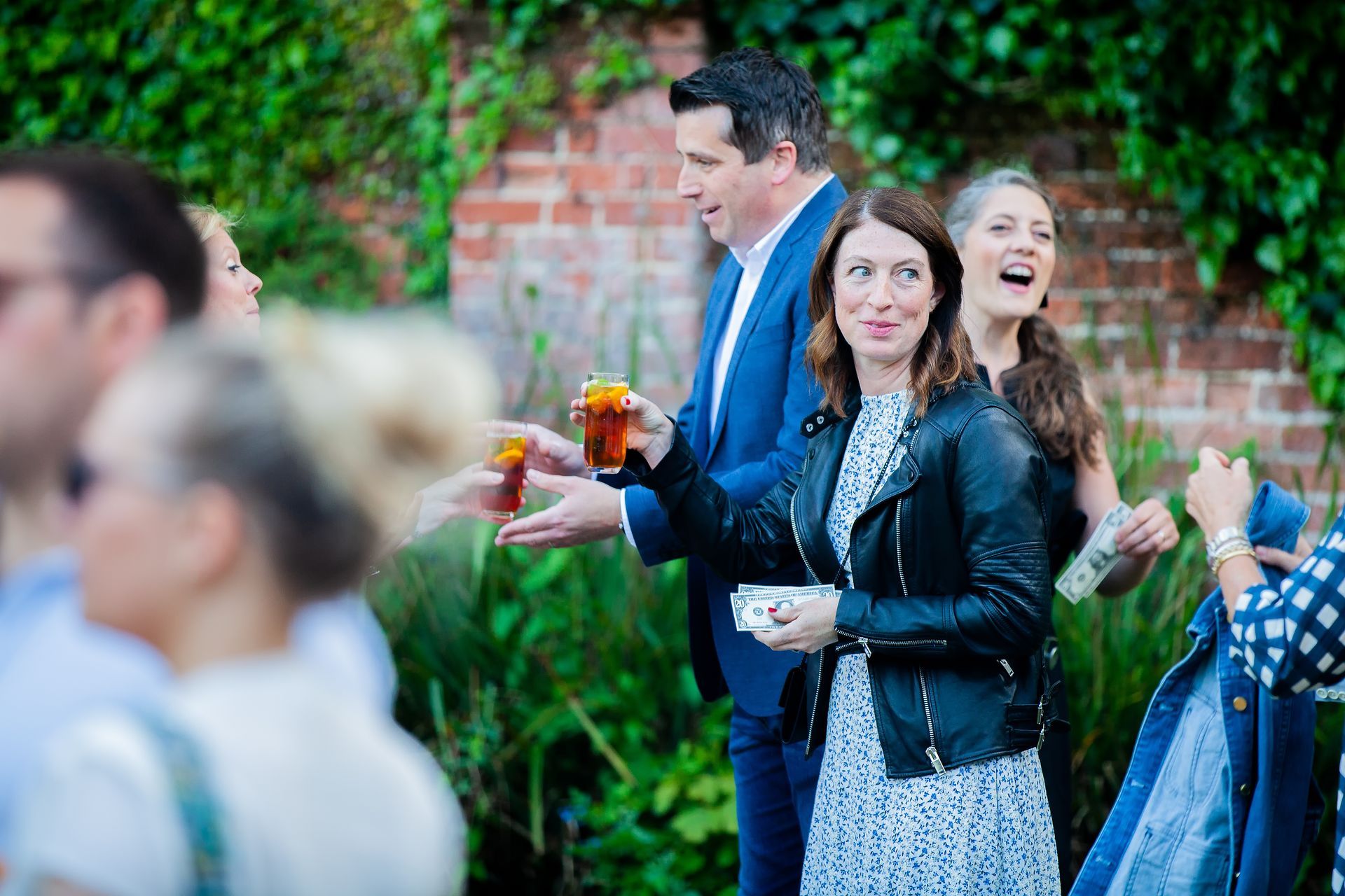 People at an outdoor event. A woman in a black jacket holds a drink; a man in a suit is behind her.