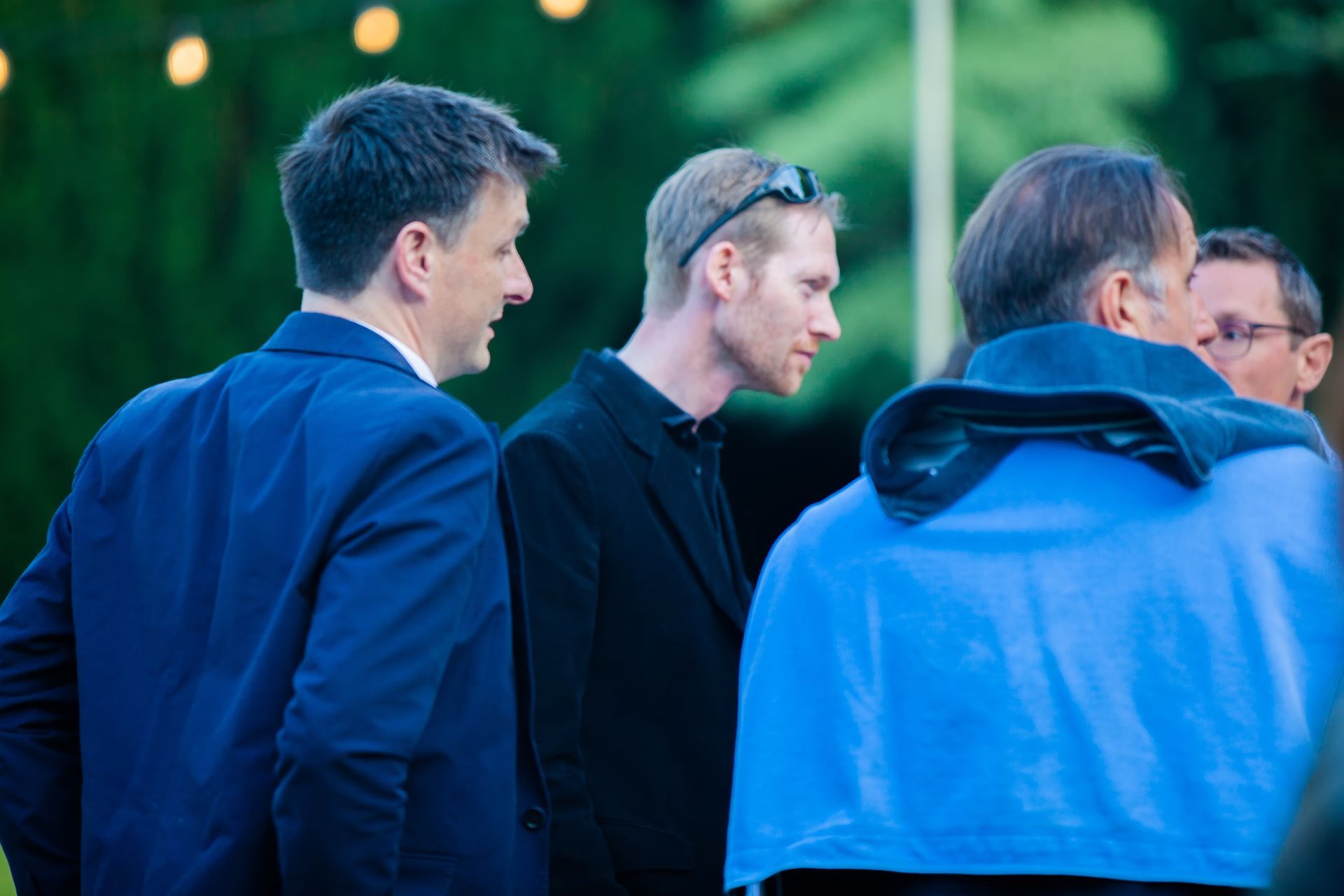 Group of men in conversation outdoors; one in blue blazer, one in black jacket, and one in a blue hoodie.