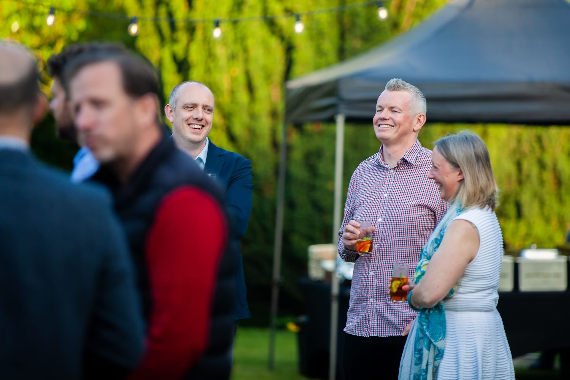 People socializing at an outdoor event; some are smiling and holding drinks. Green trees and a dark tent are in the background.