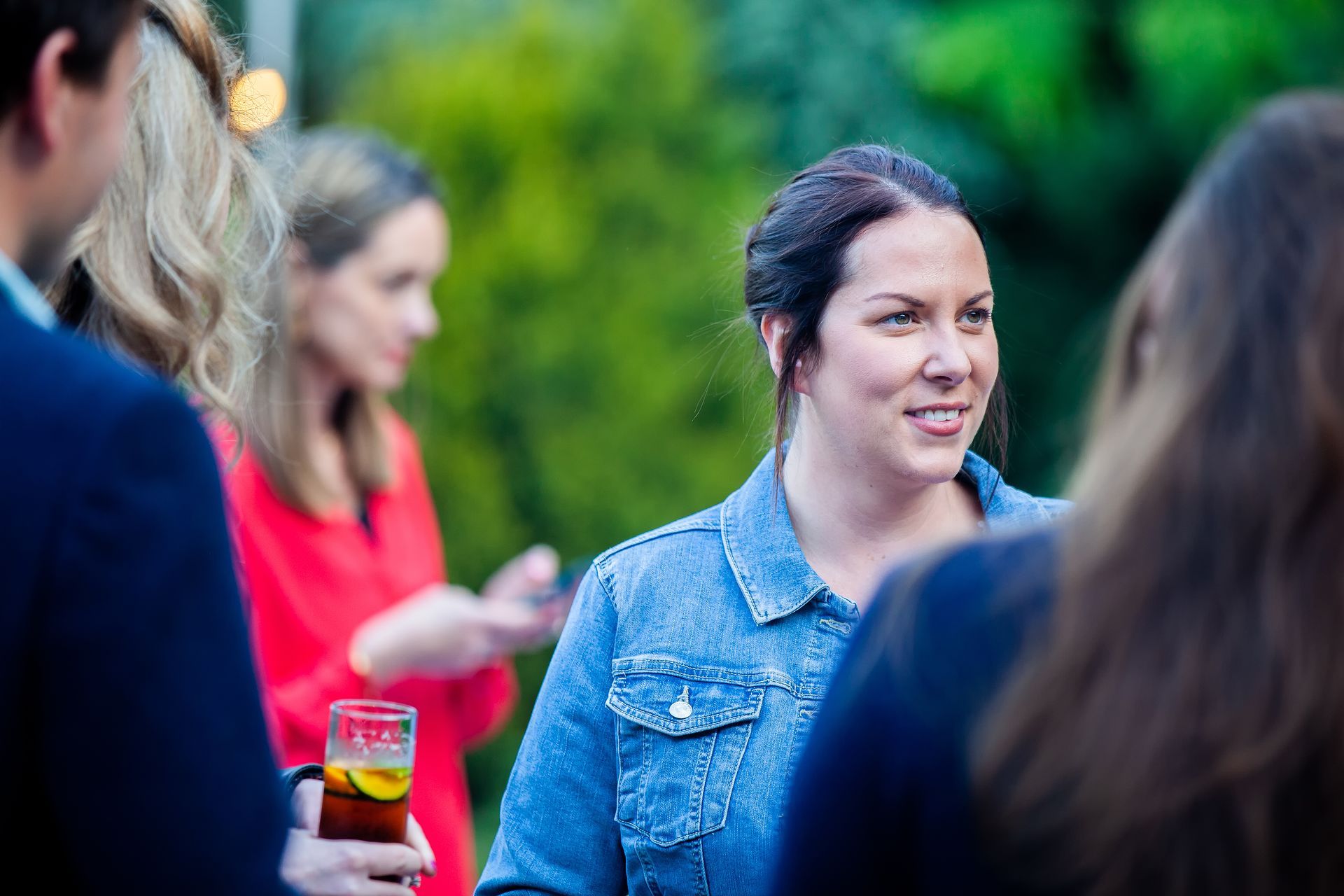 Woman in denim jacket smiles, talking with others outdoors at an event; red drink in hand.