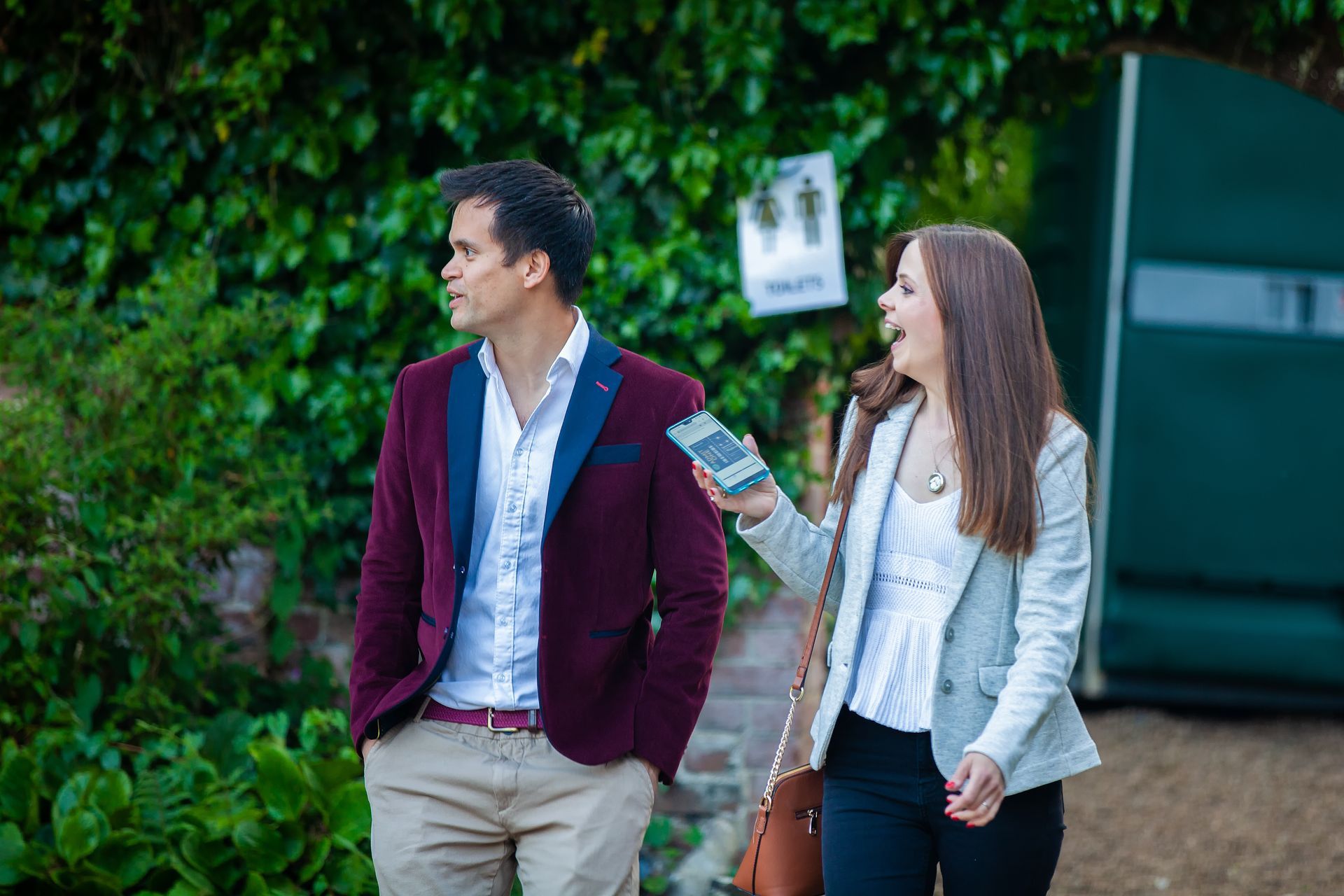 Man and woman walk near a restroom sign; man looks away, woman holds phone, both in semi-formal wear.