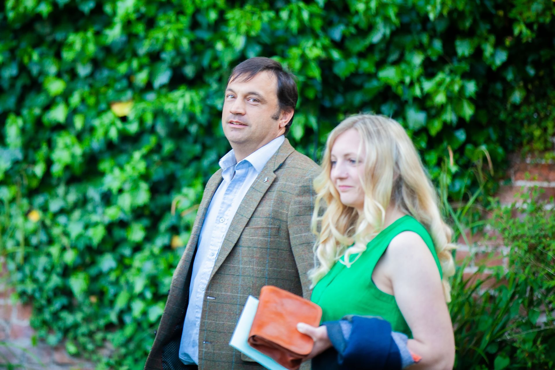Man and woman walking, man in tweed jacket, woman in green dress, brick wall with vines.