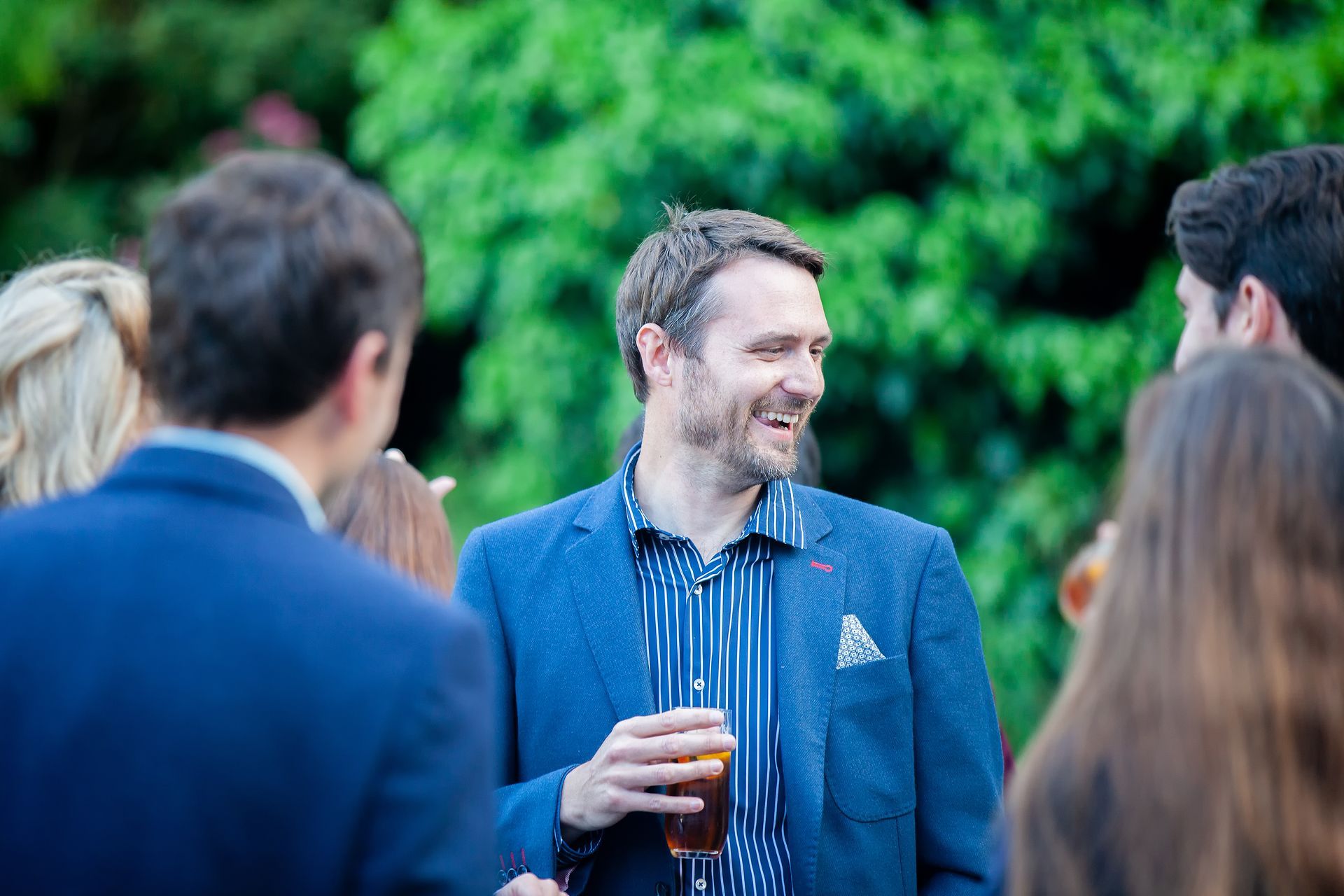 Man in blue blazer smiles, holding a drink, talking with a group outdoors.