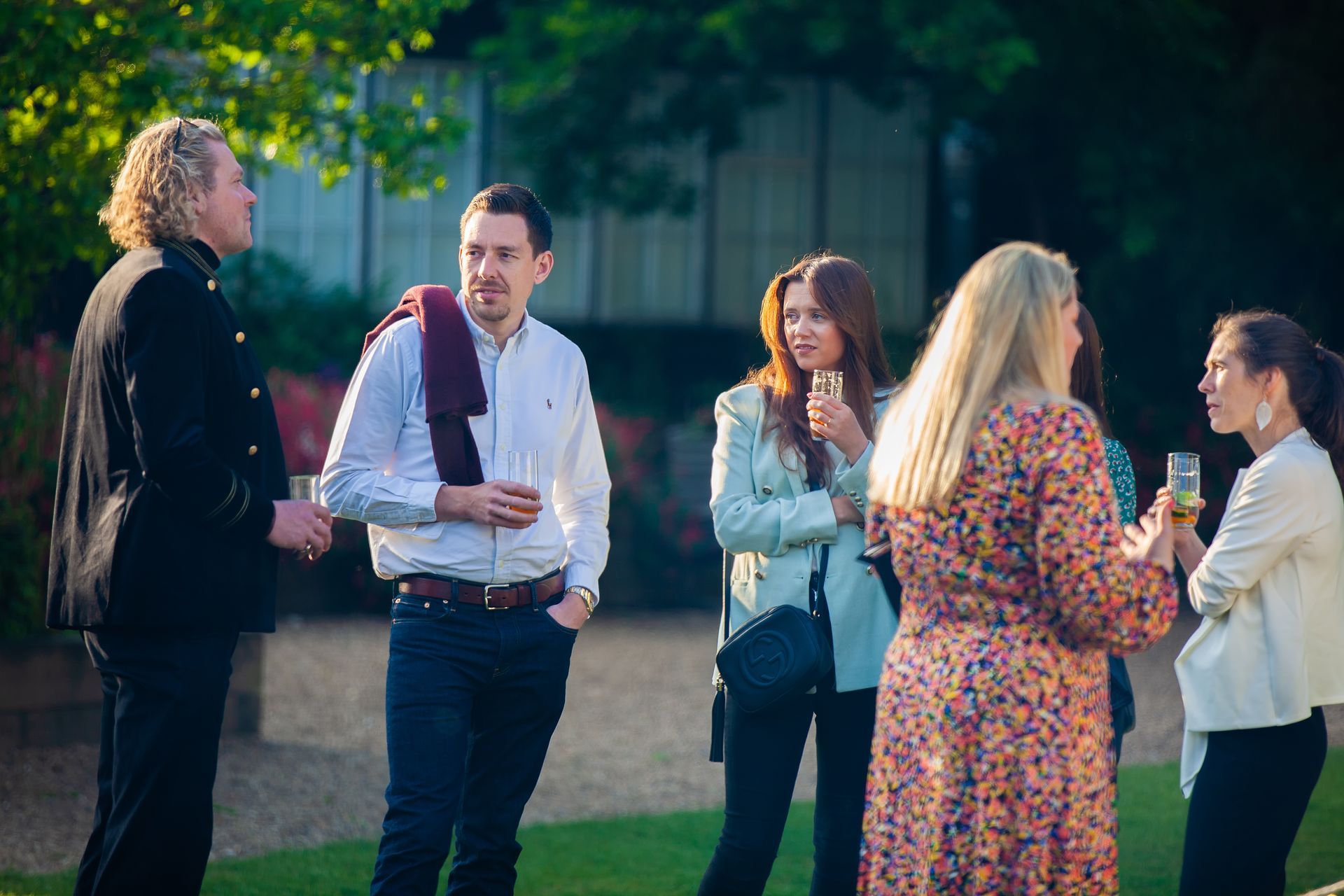 Group of people socializing outdoors, holding drinks. Men in suits, others casual, talking in garden.