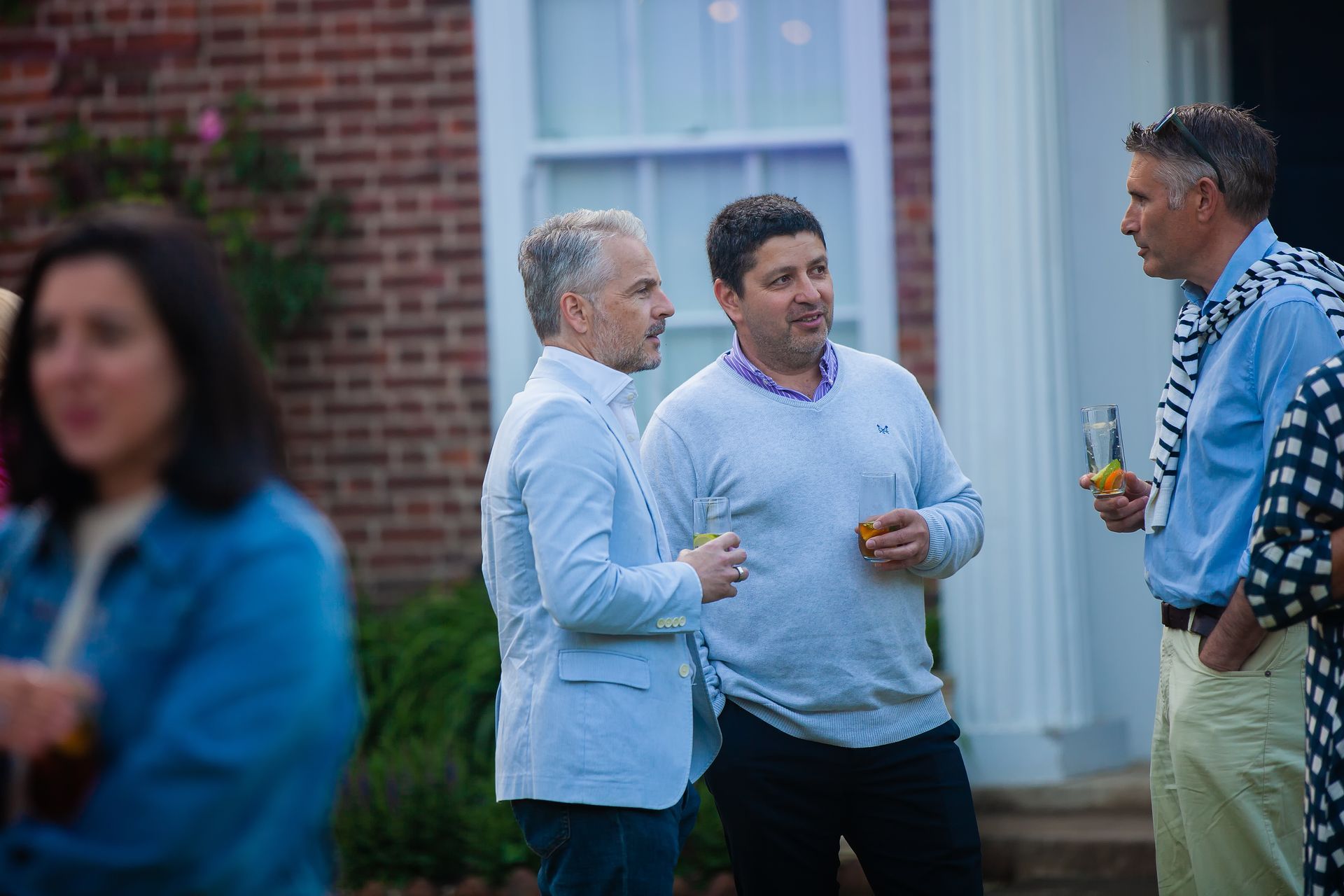 Three men converse outdoors with drinks, near a brick building with white columns.