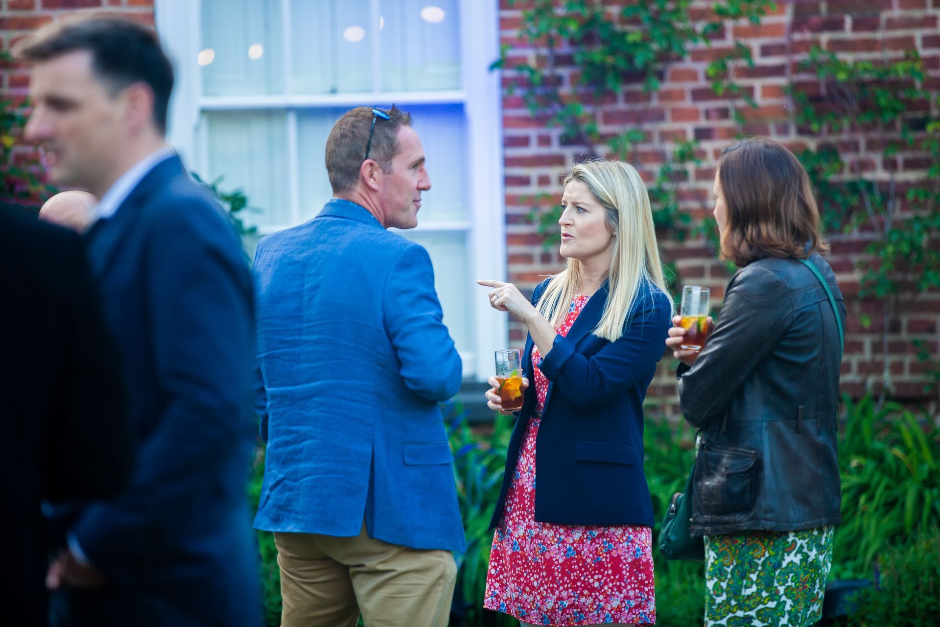 People in a garden, chatting and holding drinks. A blonde woman points while speaking. Brick building in the background.