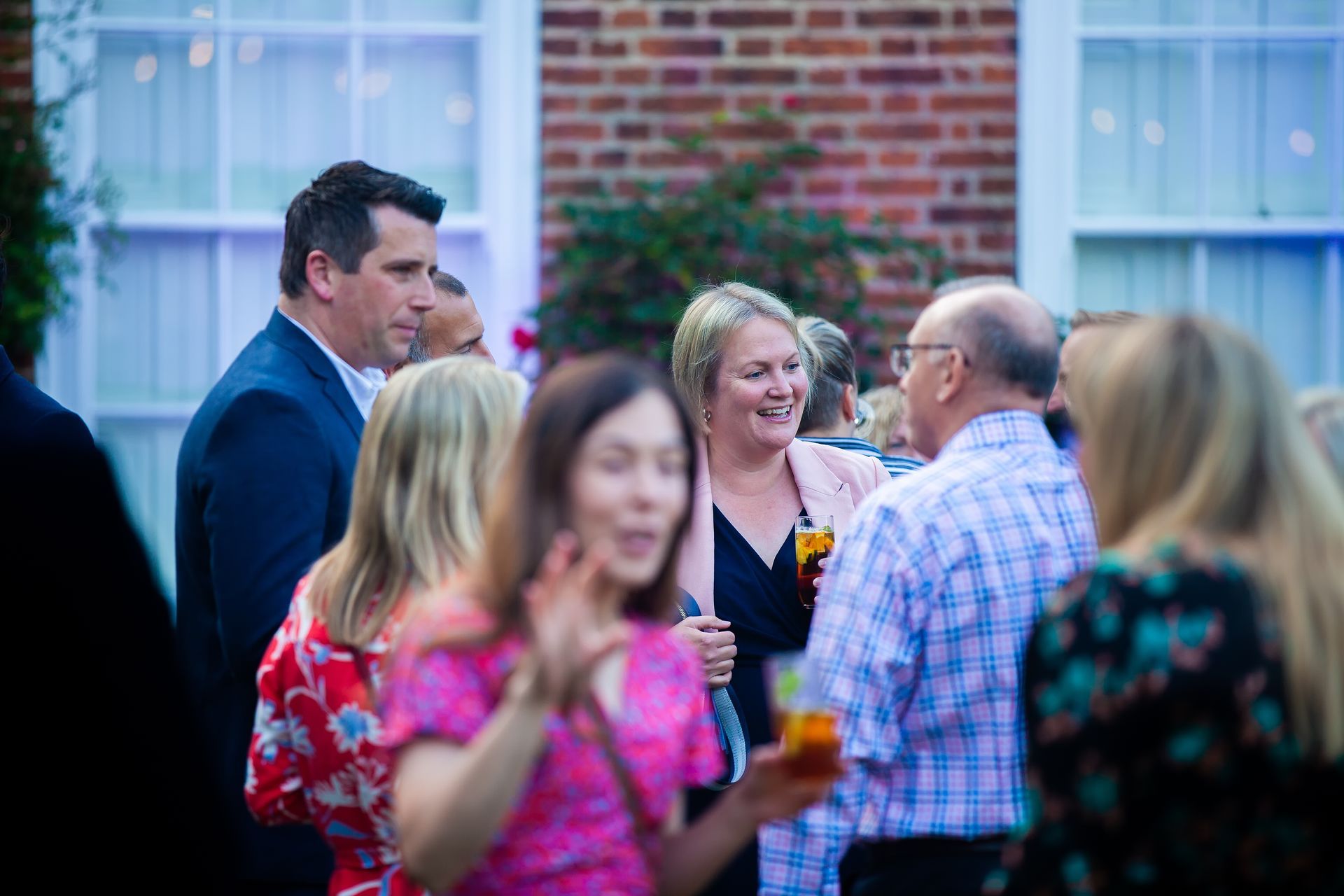 People socializing outdoors at an event. Woman smiles, drinks in hand. Man in blue suit. Brick building background.