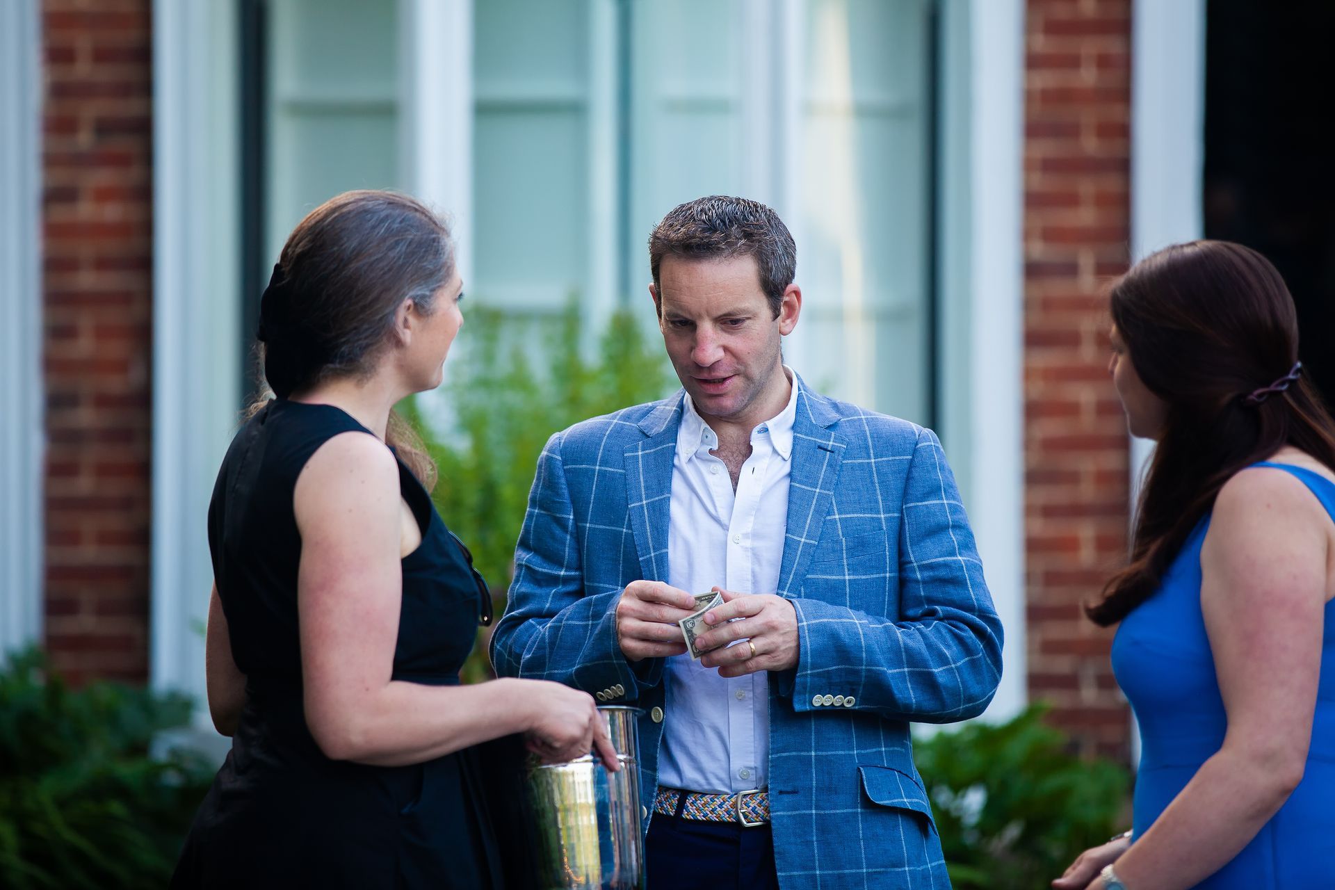 Three people conversing outside a brick building. The man in a blue blazer holds cash. Women wear black and blue dresses.