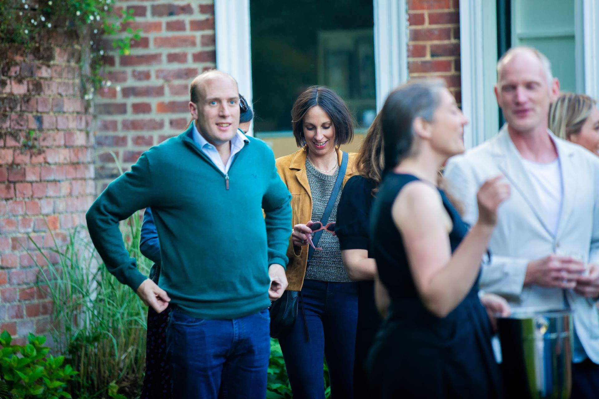 Man in teal sweater adjusts jacket, surrounded by people at an outdoor gathering near a brick building.
