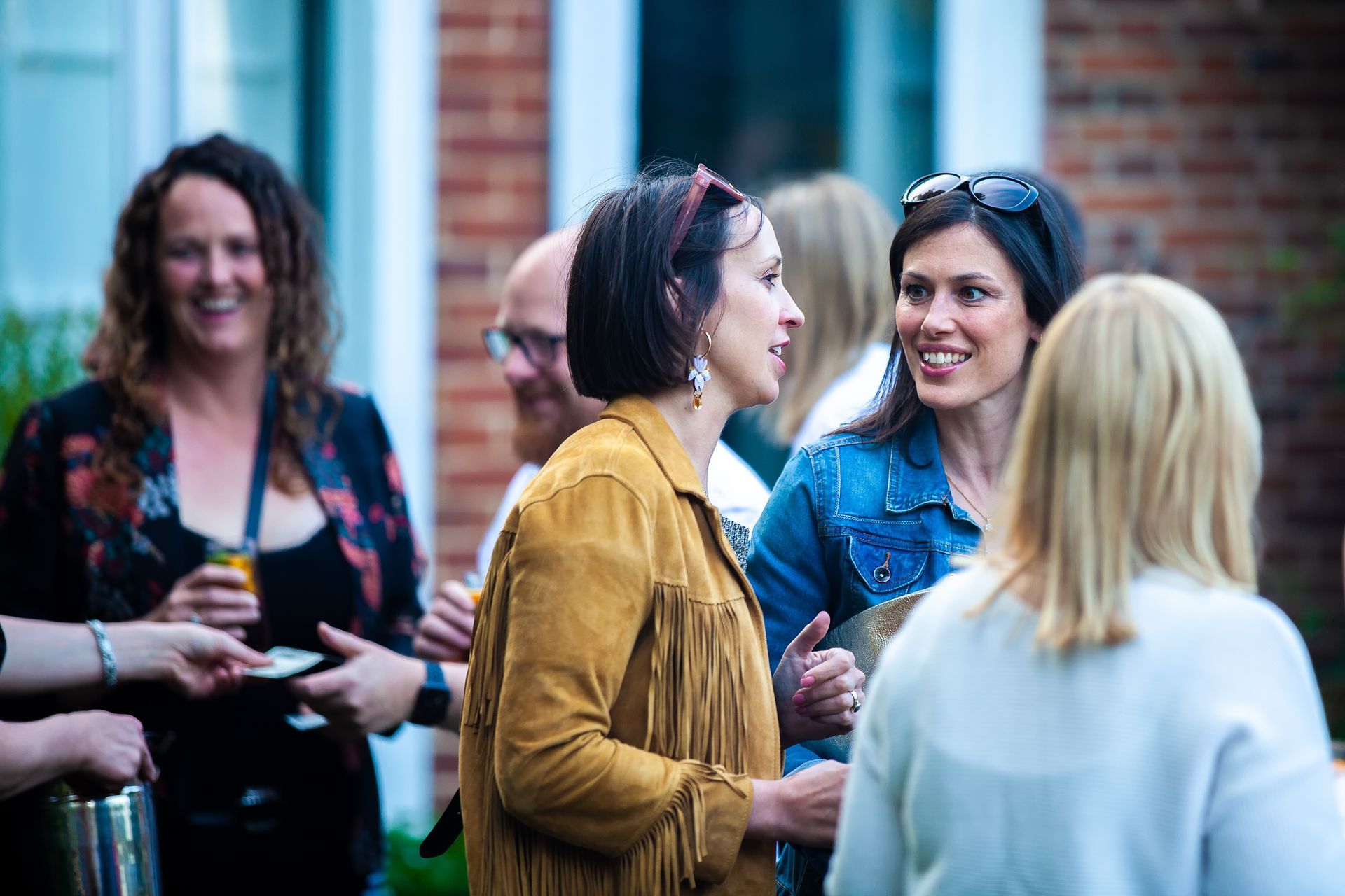 Group of women chatting outdoors, some smiling. One wears a brown jacket with fringe.