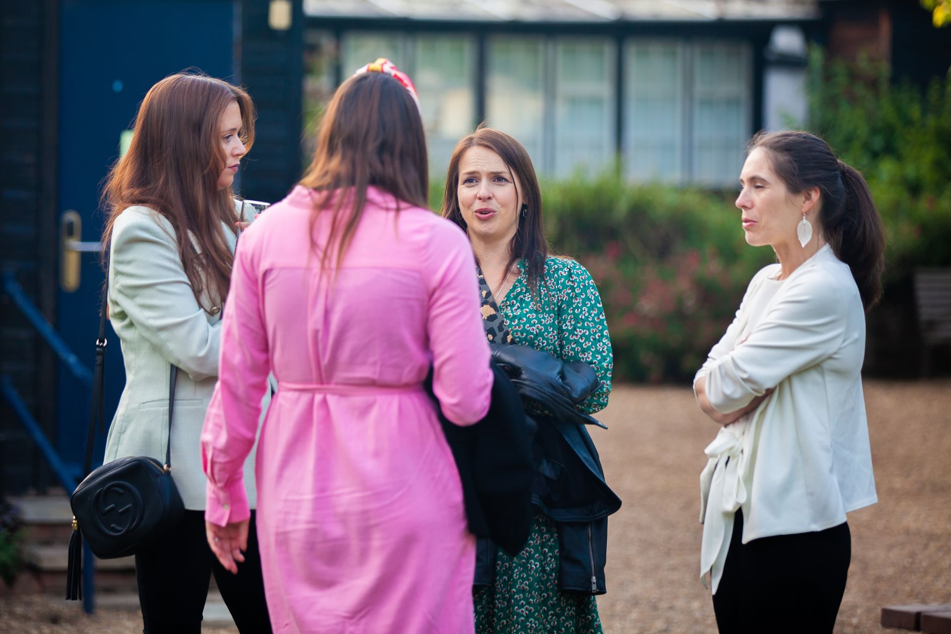 Four women conversing outdoors. One wears pink, others wear white jackets, one in green dress. Daytime.