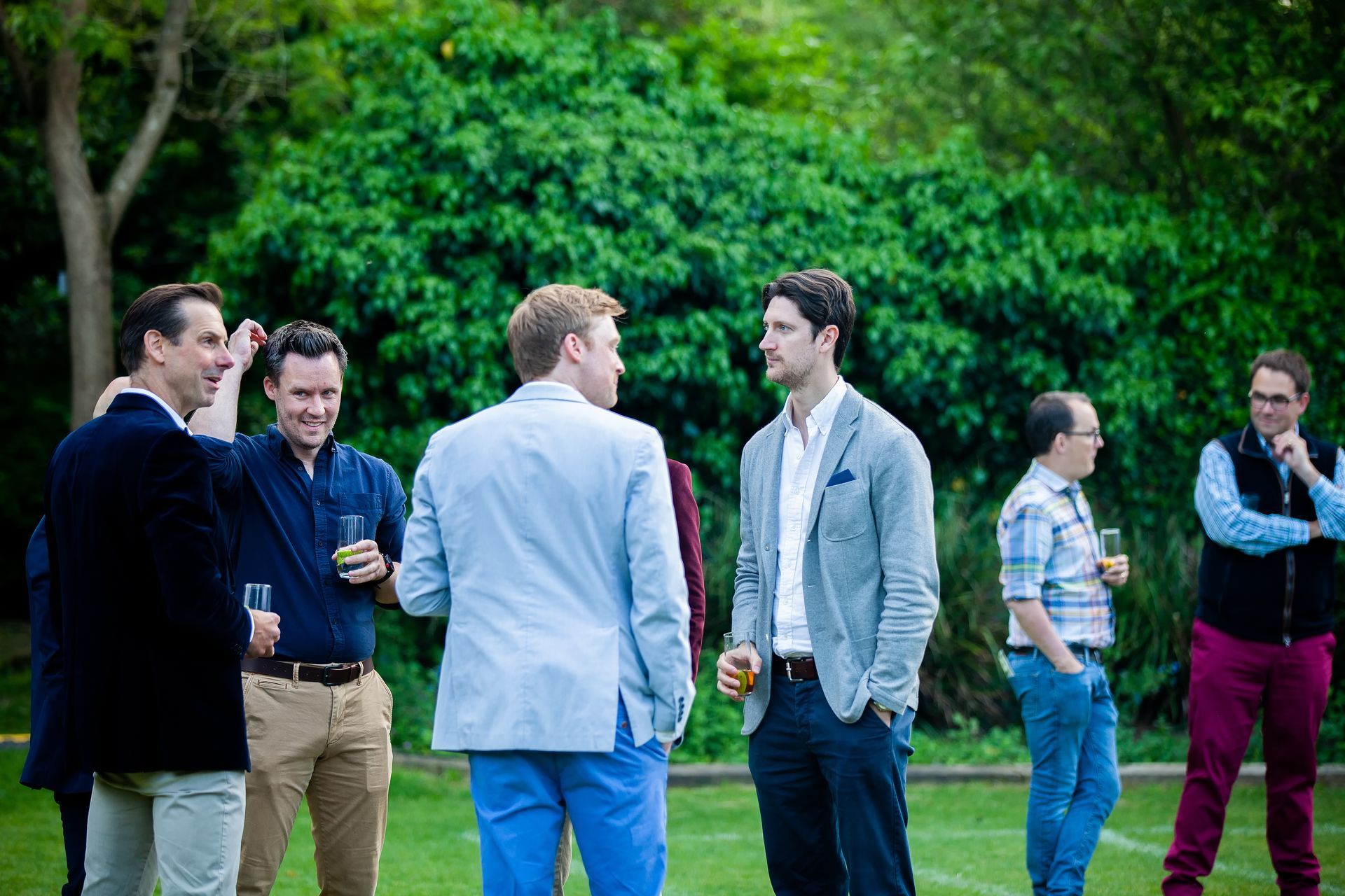 Group of men in jackets talking outdoors, holding drinks. Green grass, trees in background.