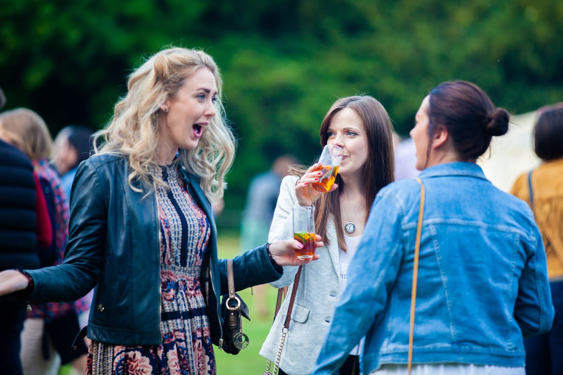 Three women at an outdoor event, chatting and drinking. One woman gestures widely, another sips a drink.