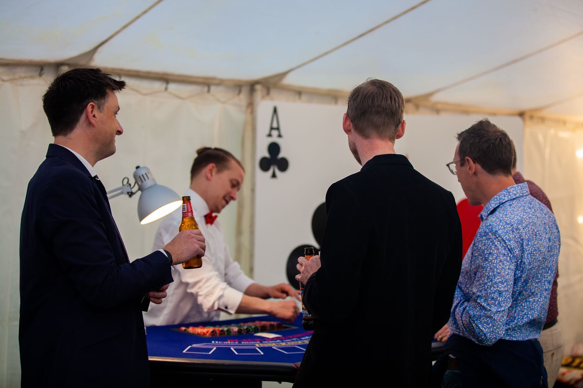 People at a casino table inside a tent. A dealer is dealing cards. One person holds a beer.