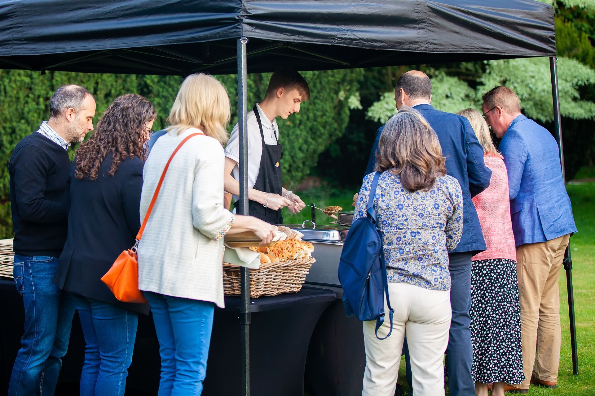 People queue at a food stall under a black canopy in a park, served by a chef.