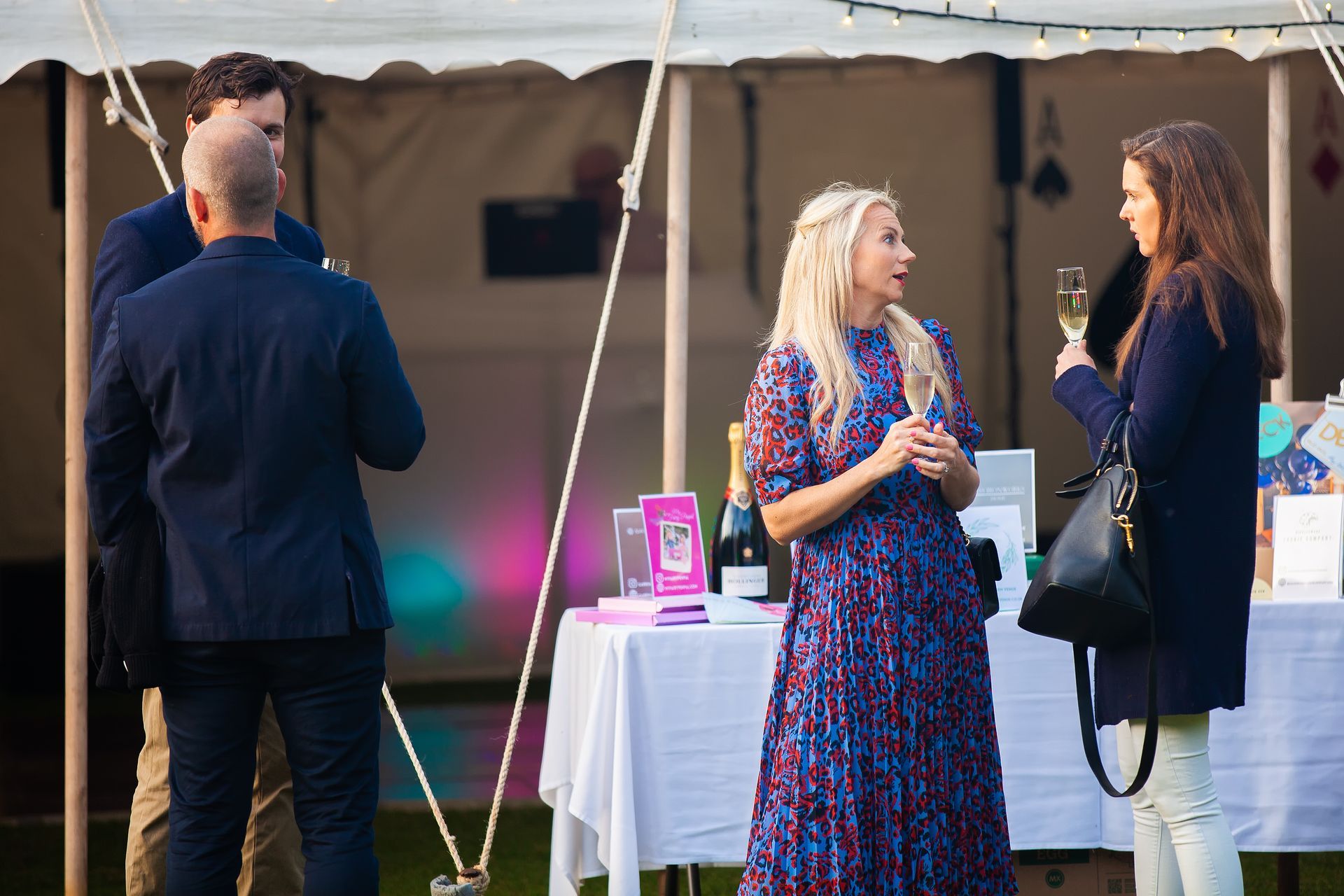 People socializing near a white tent. Woman with blonde hair in a patterned dress talks to a woman holding a glass of champagne.