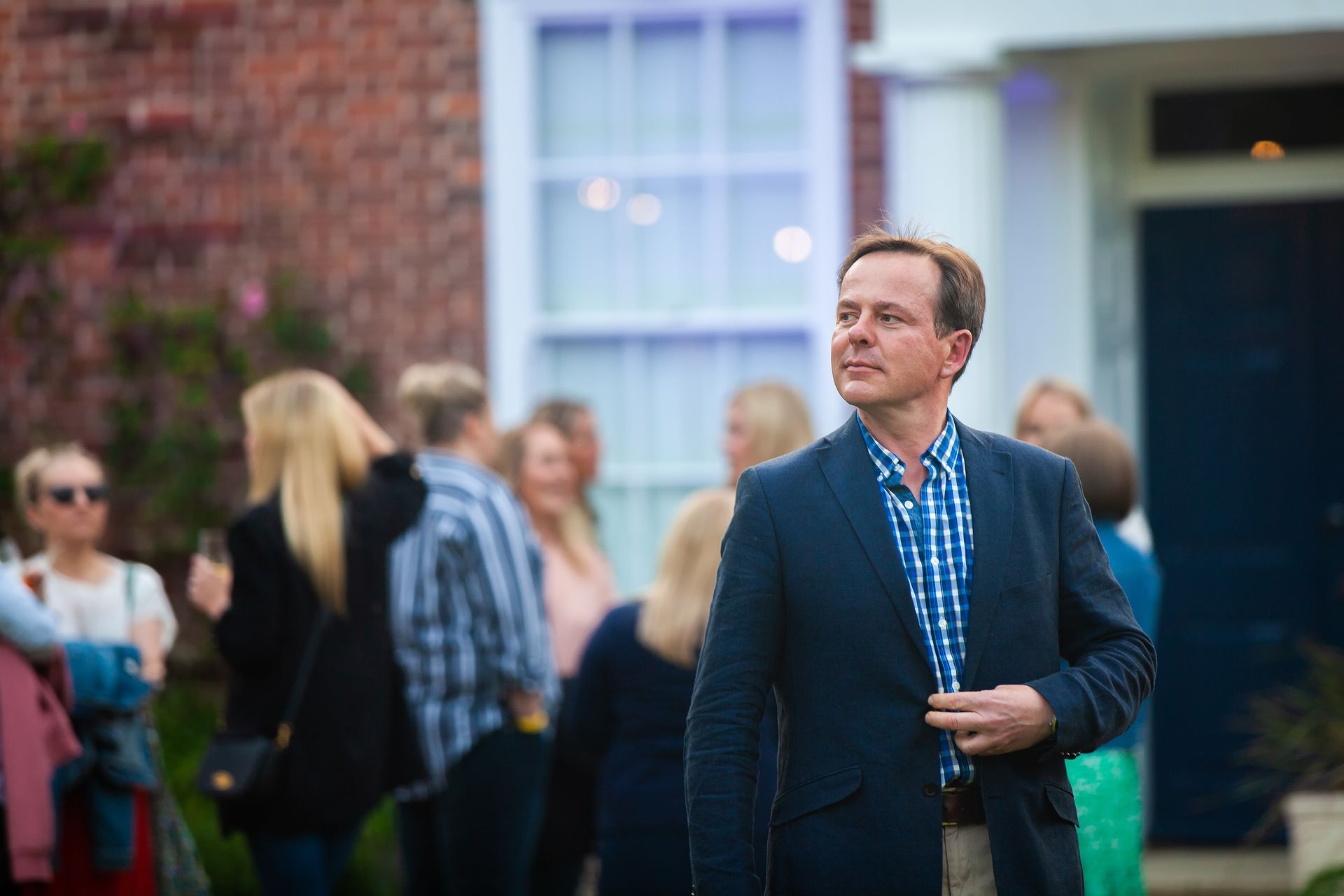Man in navy jacket at outdoor event, looking up, with others in the background near a brick building.