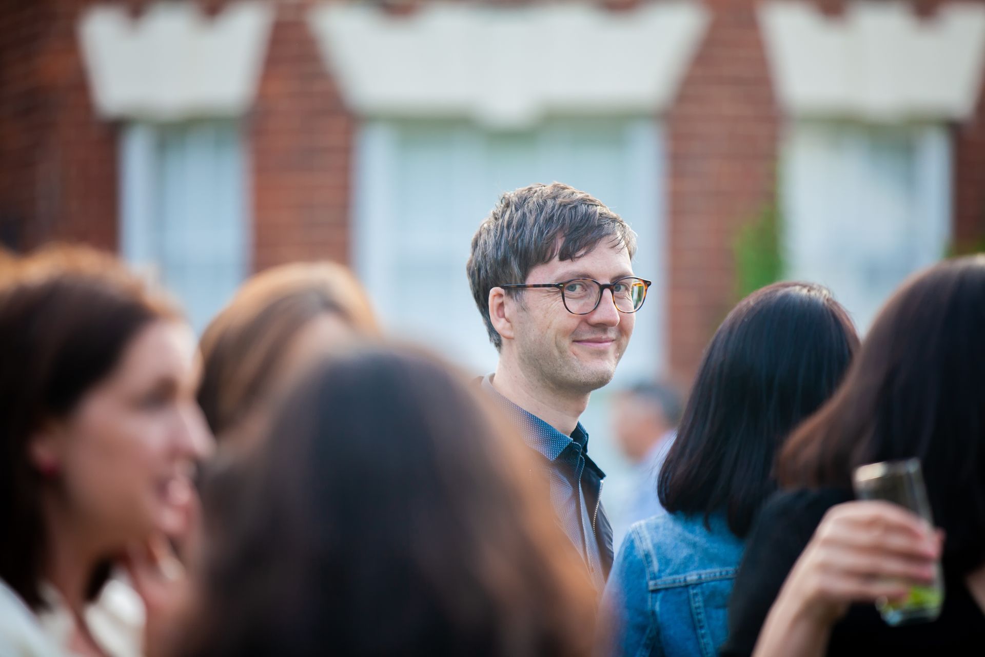 Man with glasses smiles among a crowd; brick building in background.