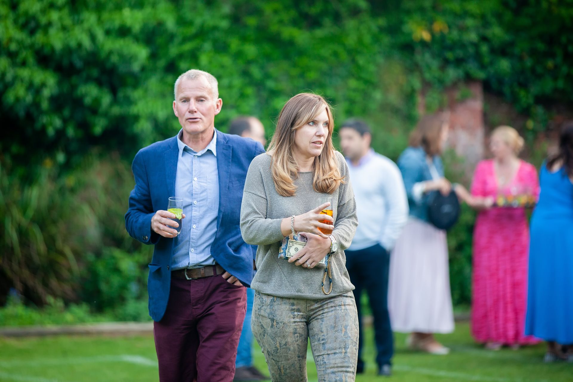 Man and woman walking on grass, drinks in hand, at an outdoor event. Other guests in background.