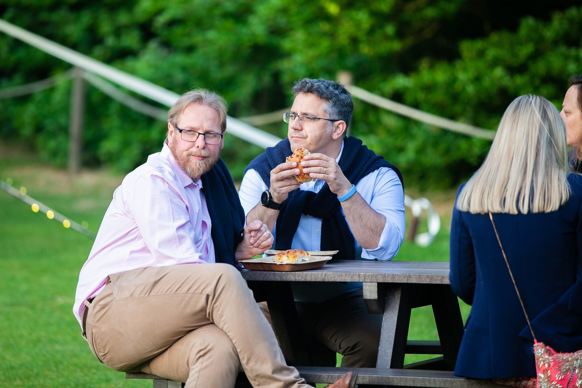 Two men eating at a picnic table outdoors. One is eating a sandwich, the other looks at the camera.