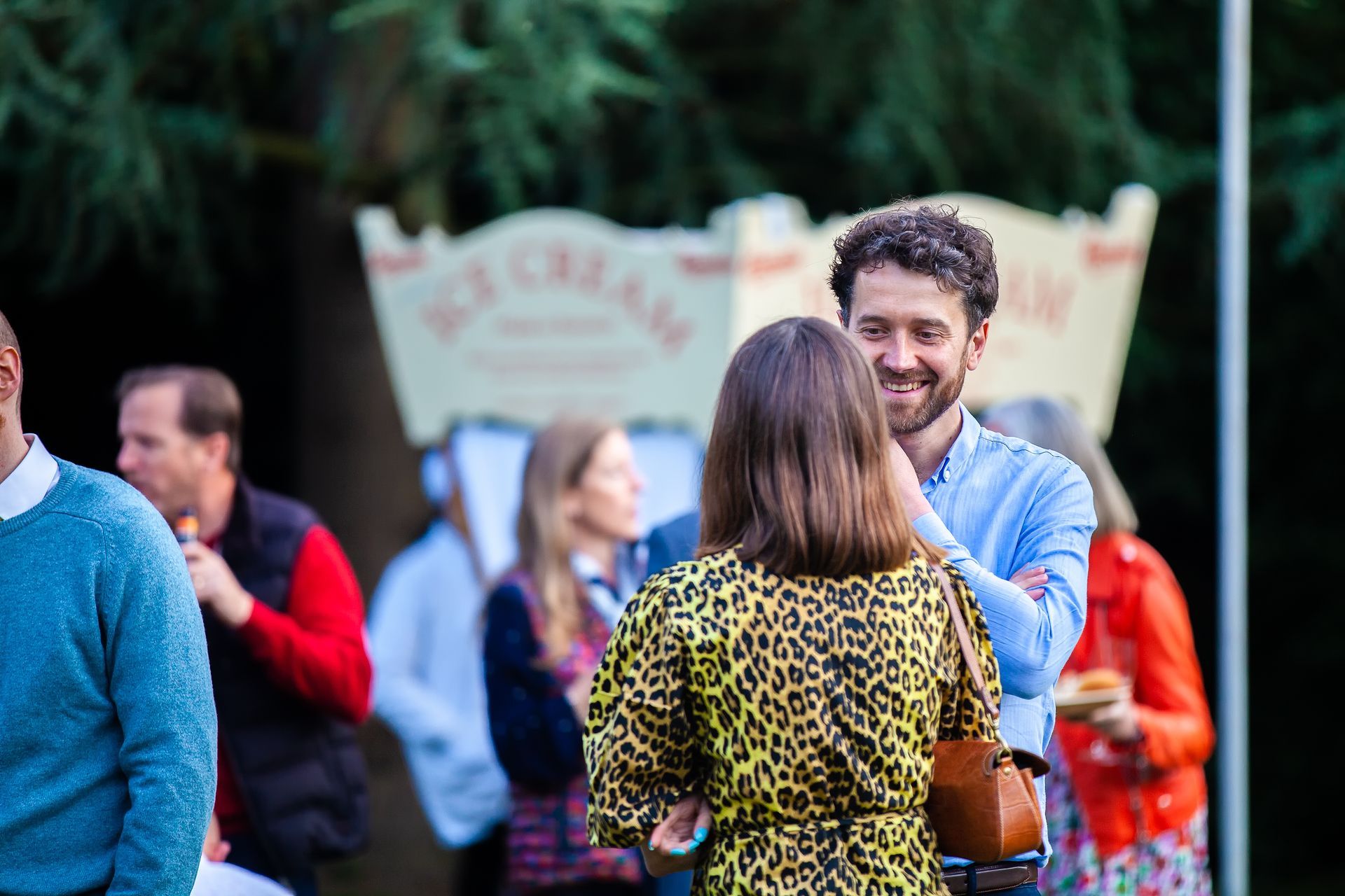 Man and woman smiling, conversing near an ice cream stand outdoors. Others in background.
