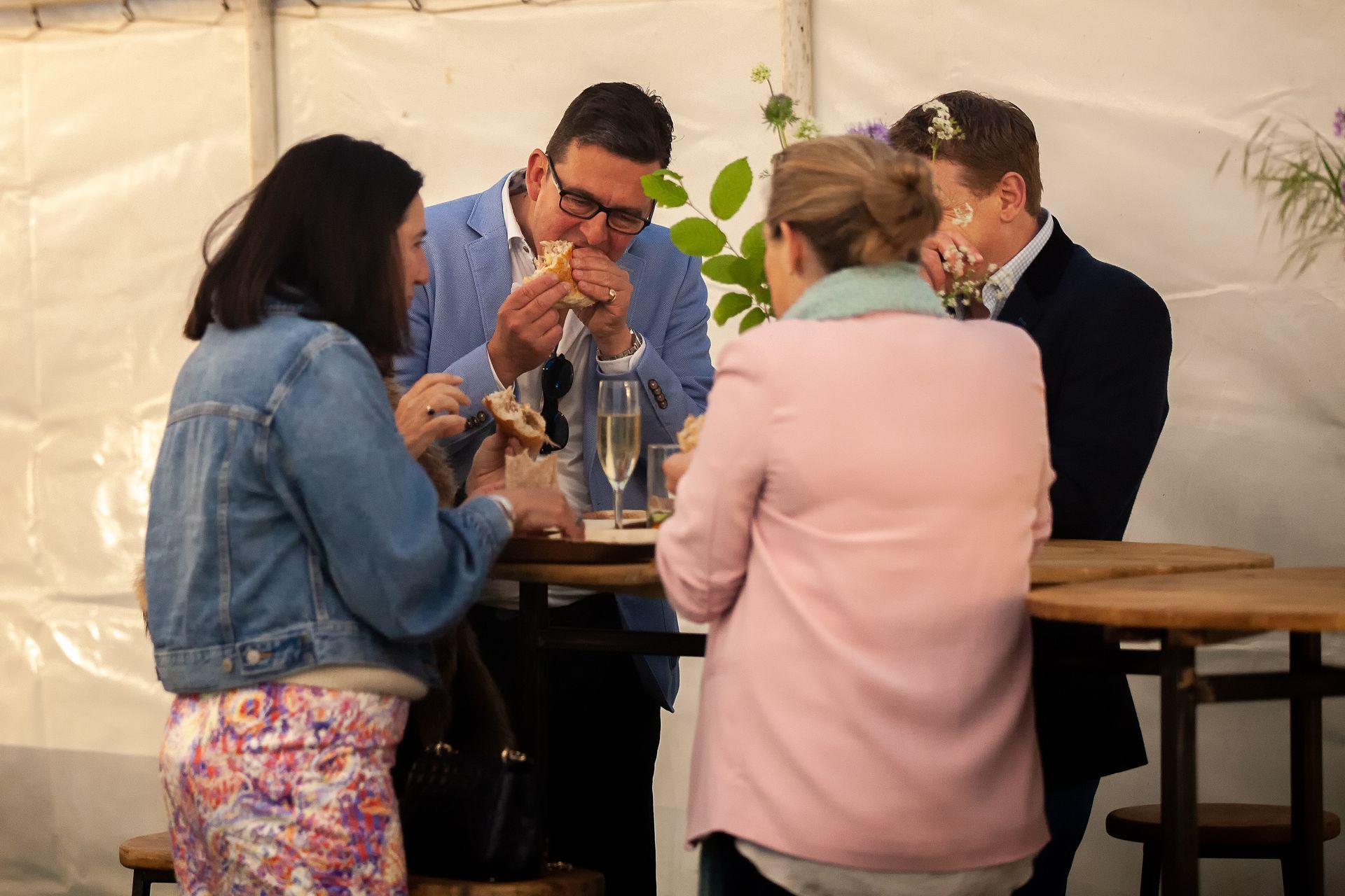 Four people eating and drinking around a table under a white tent.