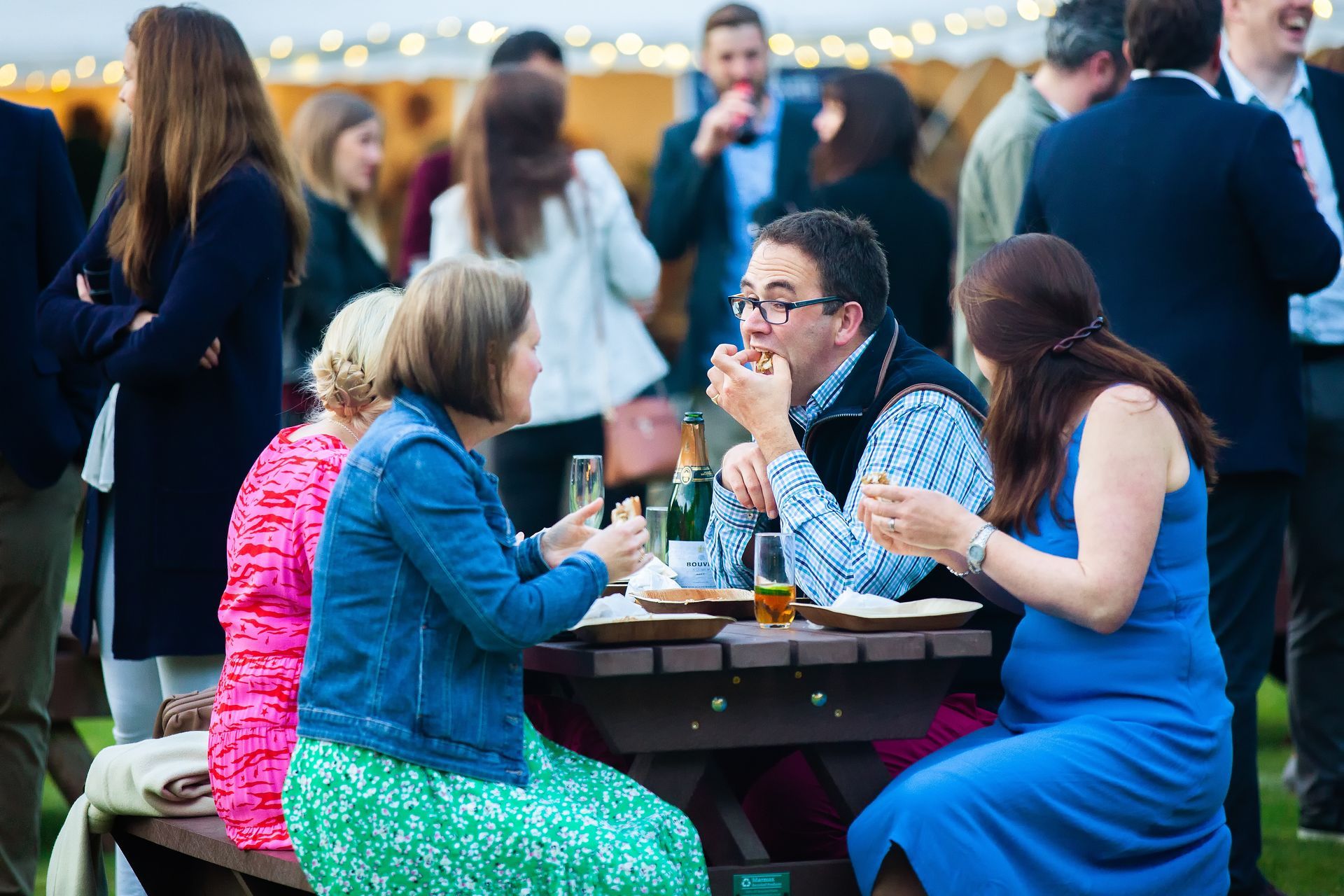 People at an outdoor event, gathered at tables, eating and talking. Many people are wearing bright colors.