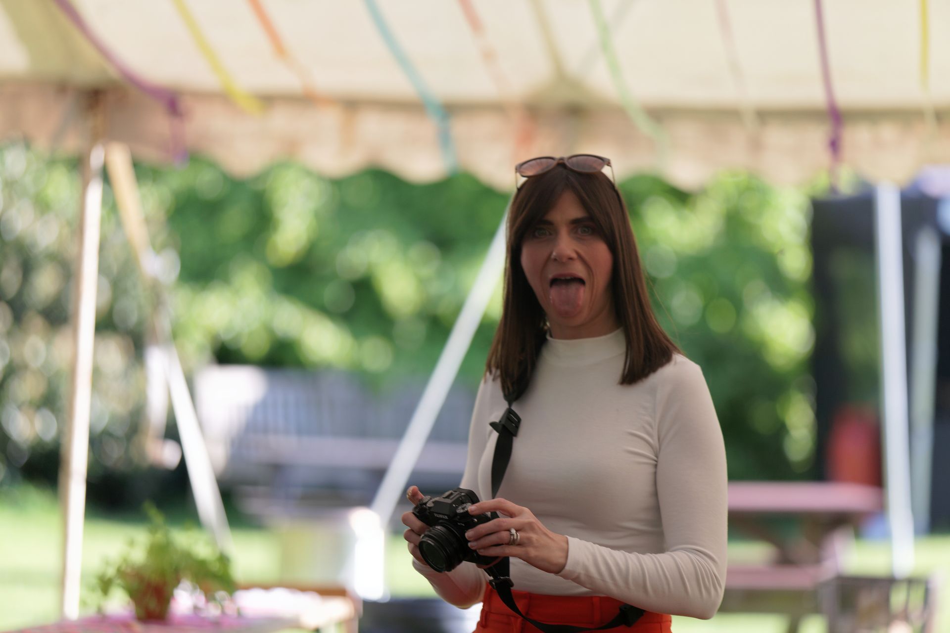 Woman sticking out her tongue, holding a camera, in a garden. She wears a white top, orange pants.