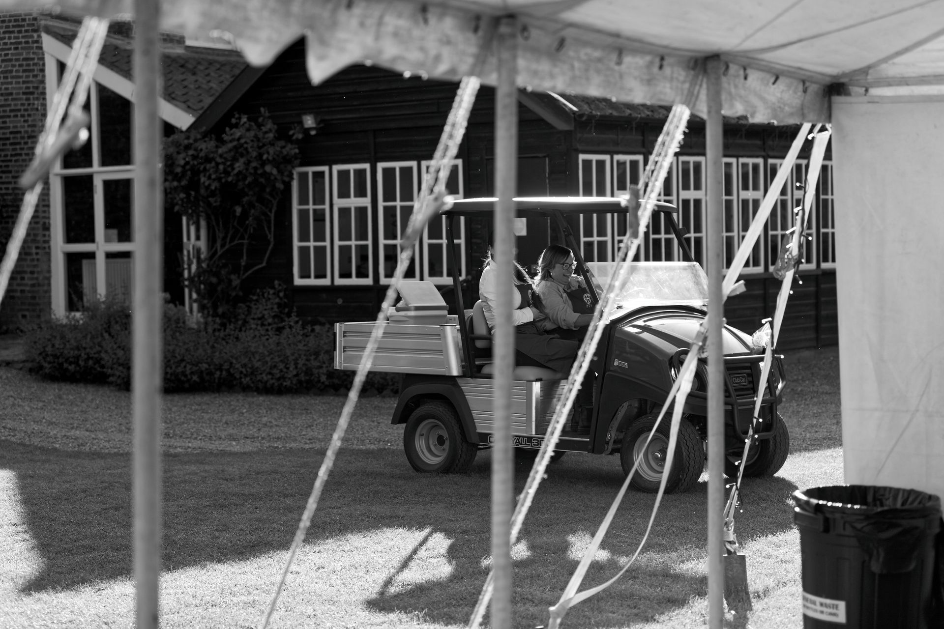 A person drives a small utility vehicle, seen through the frame of a tent. Black building in the background.