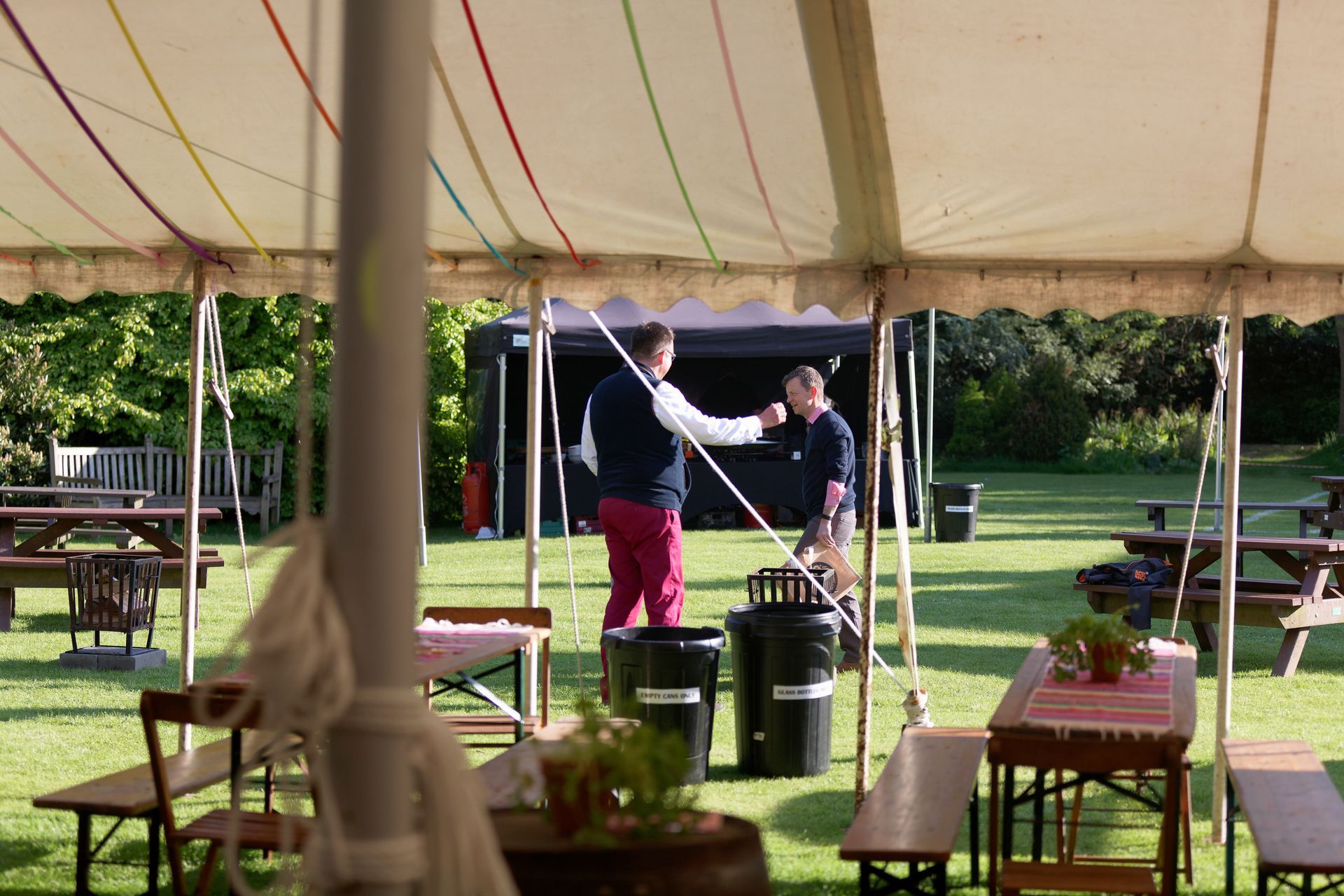 Two men under a tent, one in red pants, engaging in a discussion. Tables and benches scattered on the grass outdoors.