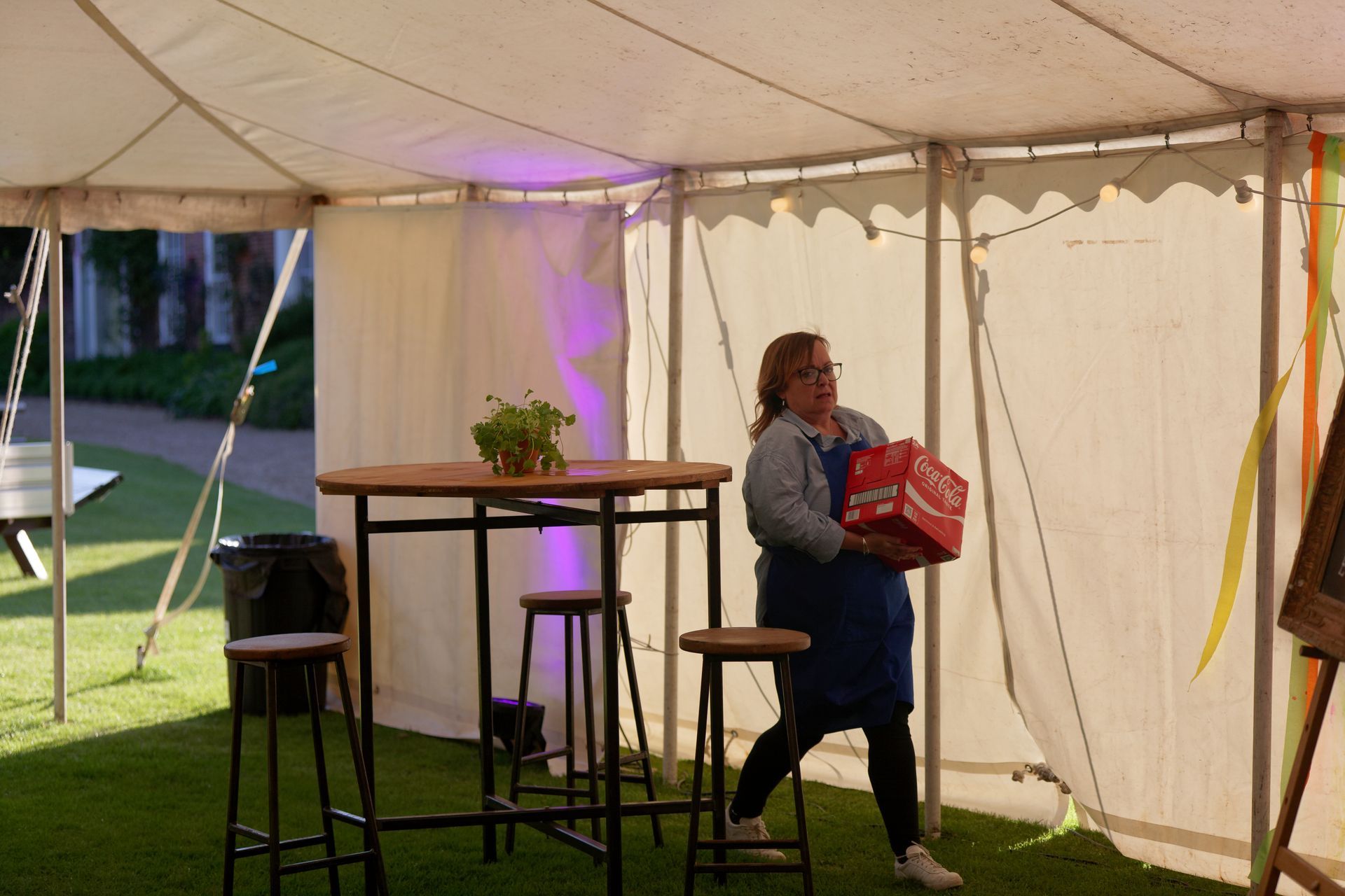 Woman carries a case of drinks inside a tent, past a table and stools on a lawn.
