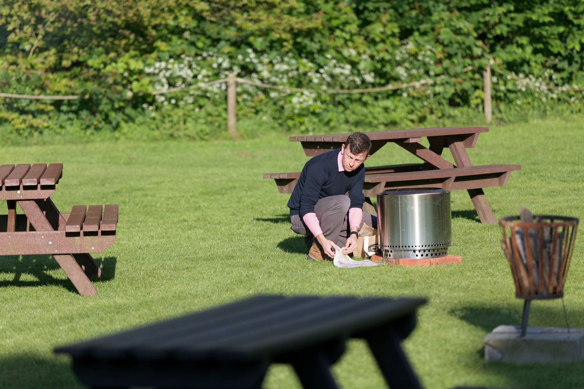 Man kneeling near a fire pit in a grassy area with picnic tables and trees in the background.