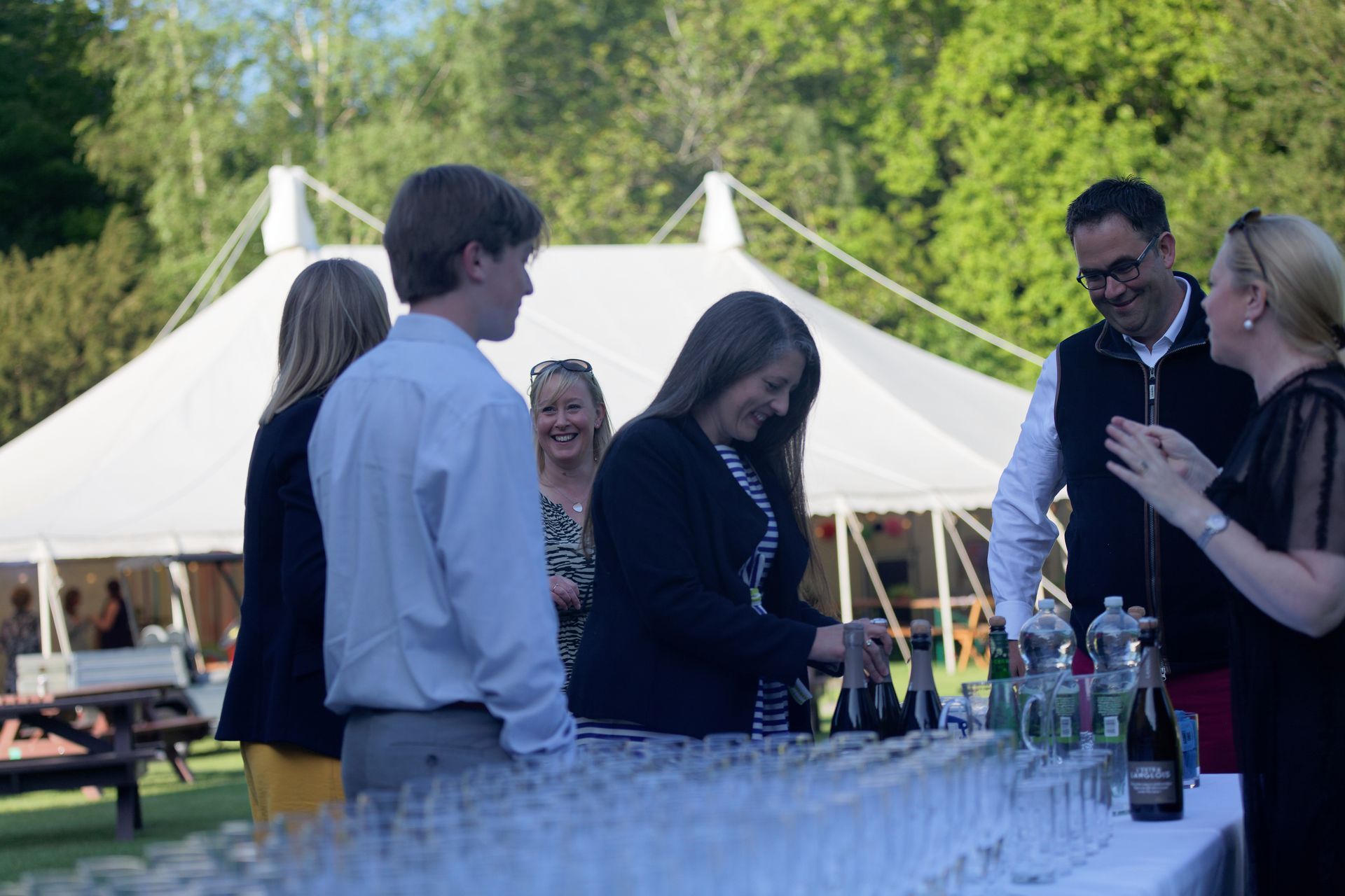 People at an outdoor event. A woman pours champagne into glasses on a table. A white tent is in the background.