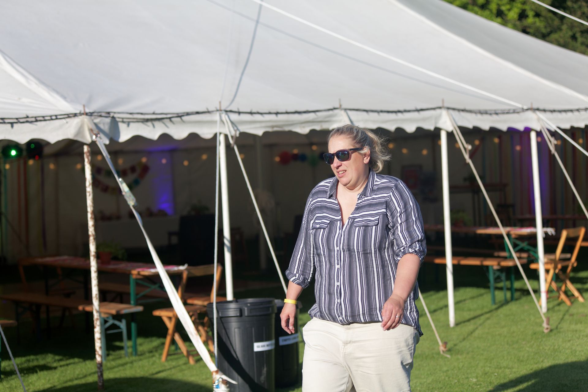 Woman in sunglasses and patterned shirt standing near a white tent with tables and a grassy lawn.