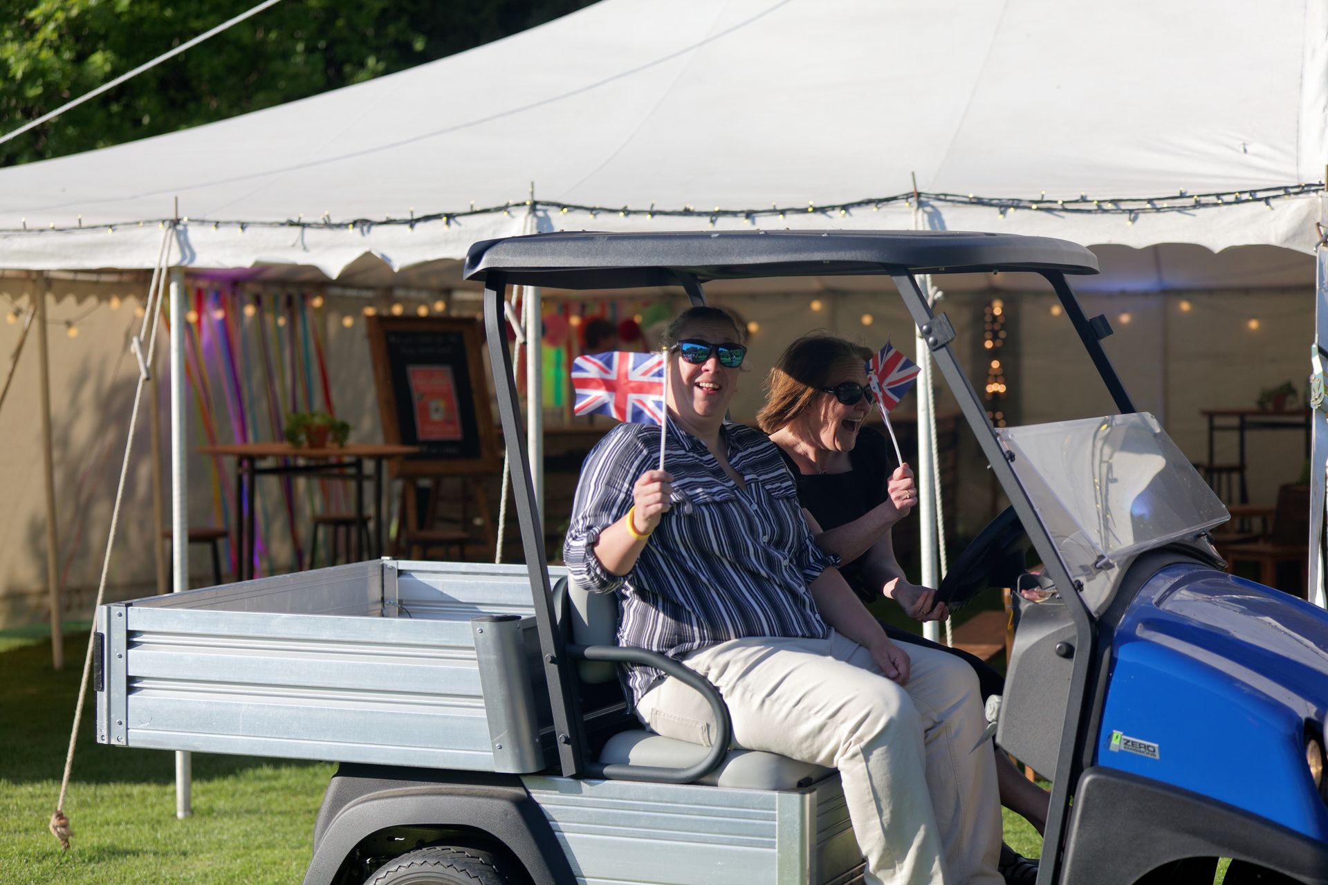 Two people in a blue golf cart, waving Union Jack flags, in front of a white tent.