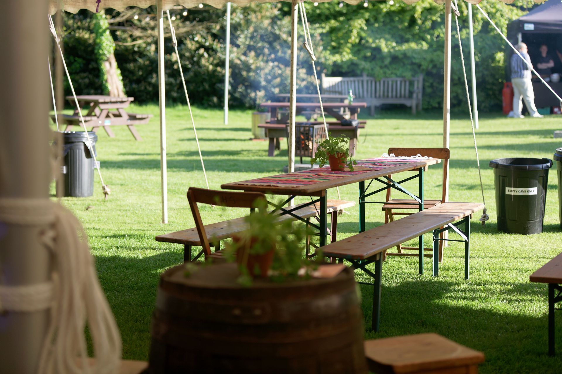 Outdoor event: Tables set under tent on green grass, with benches, and a barrel in the foreground.
