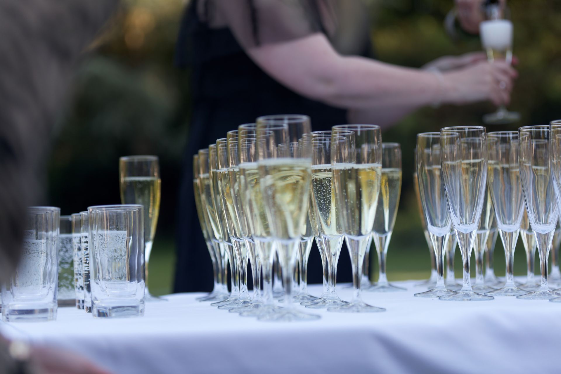 Rows of champagne flutes on a table, being filled by a person in the background.