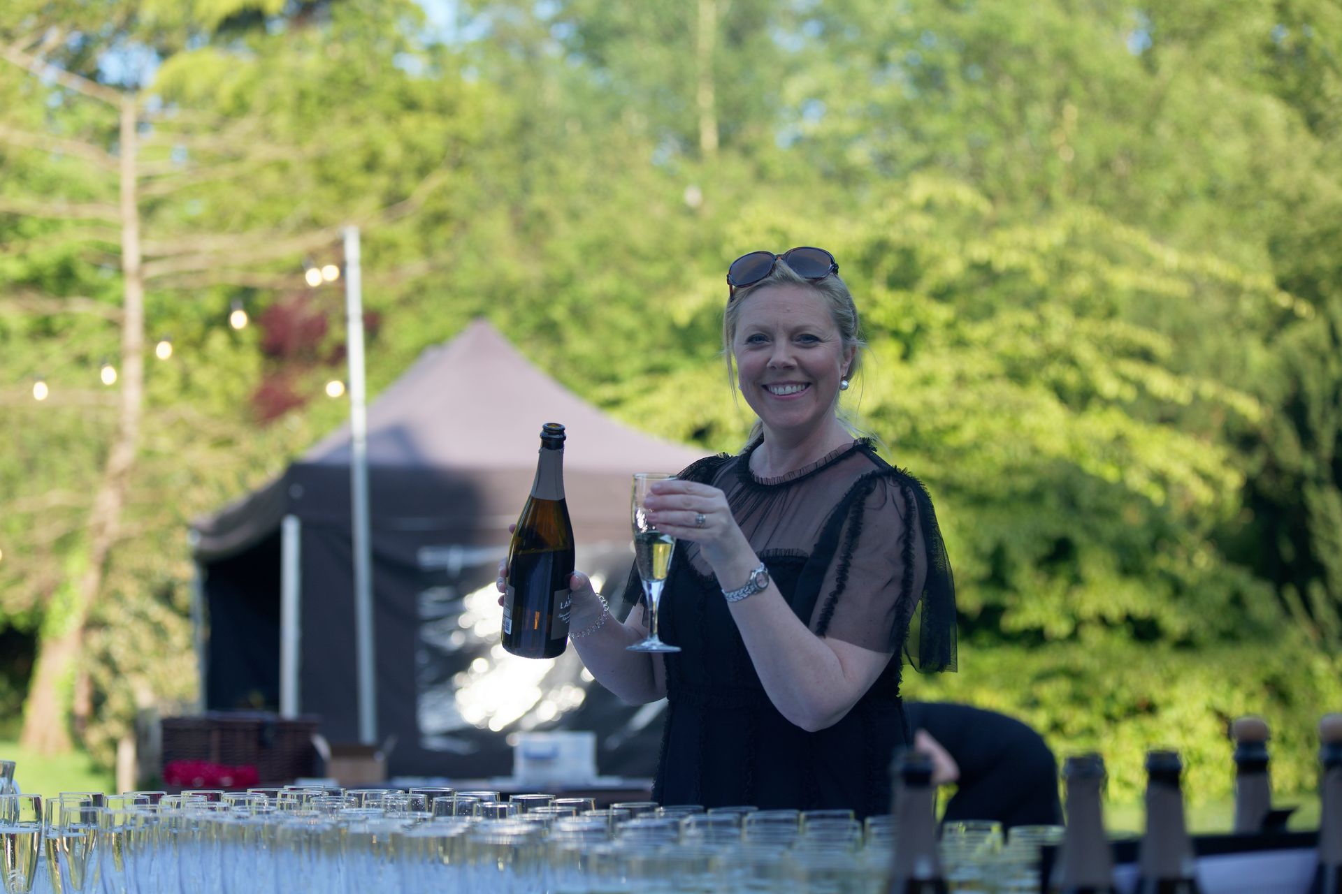 Woman pouring champagne at an outdoor bar, smiling. She wears black in a park-like setting.