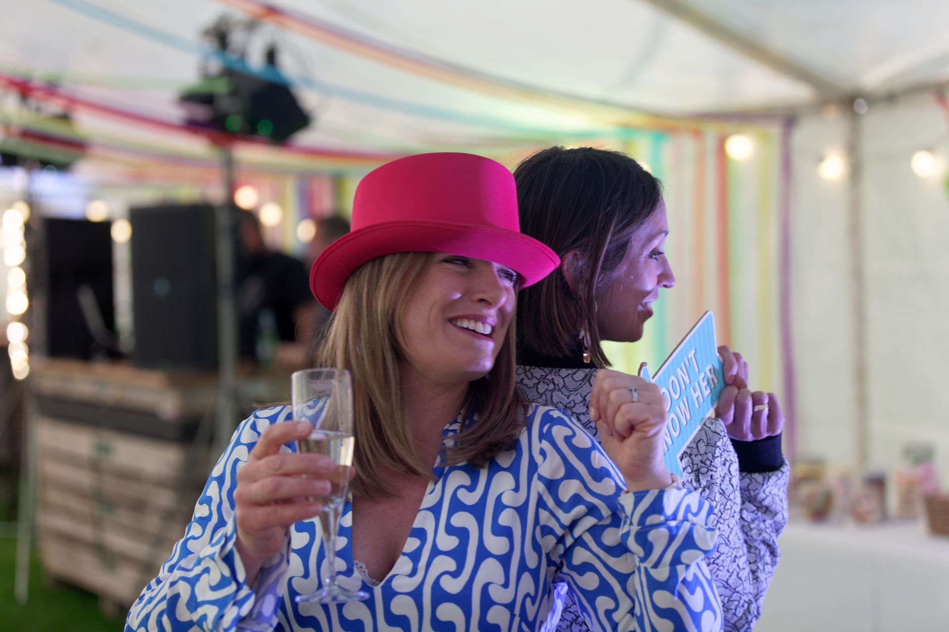 Two women at a party; one in a pink hat and blue patterned outfit, holding champagne, smiling.