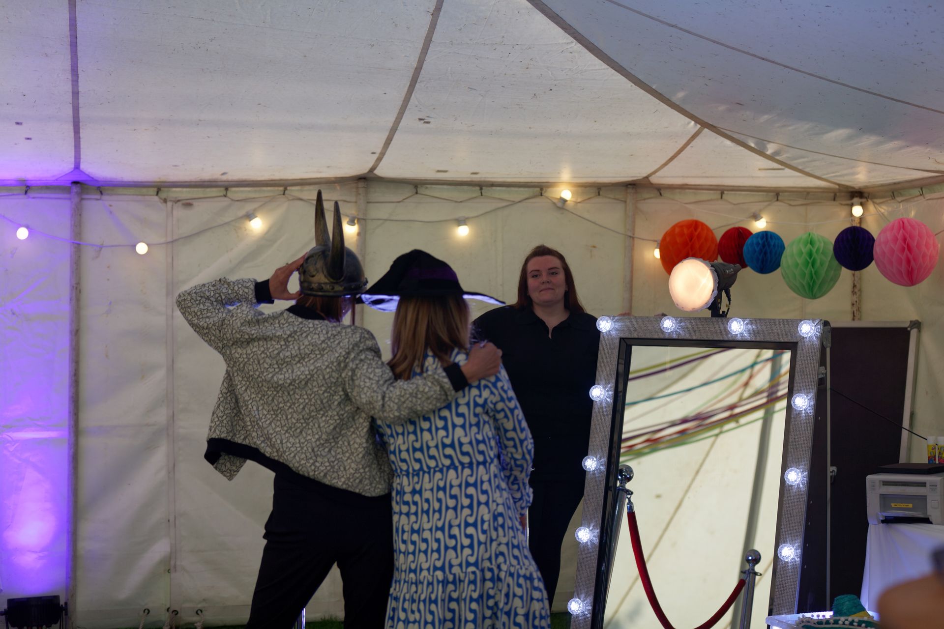 Two people pose in front of a photo booth mirror, one wearing a hat. A third person stands behind them. Colorful decorations.
