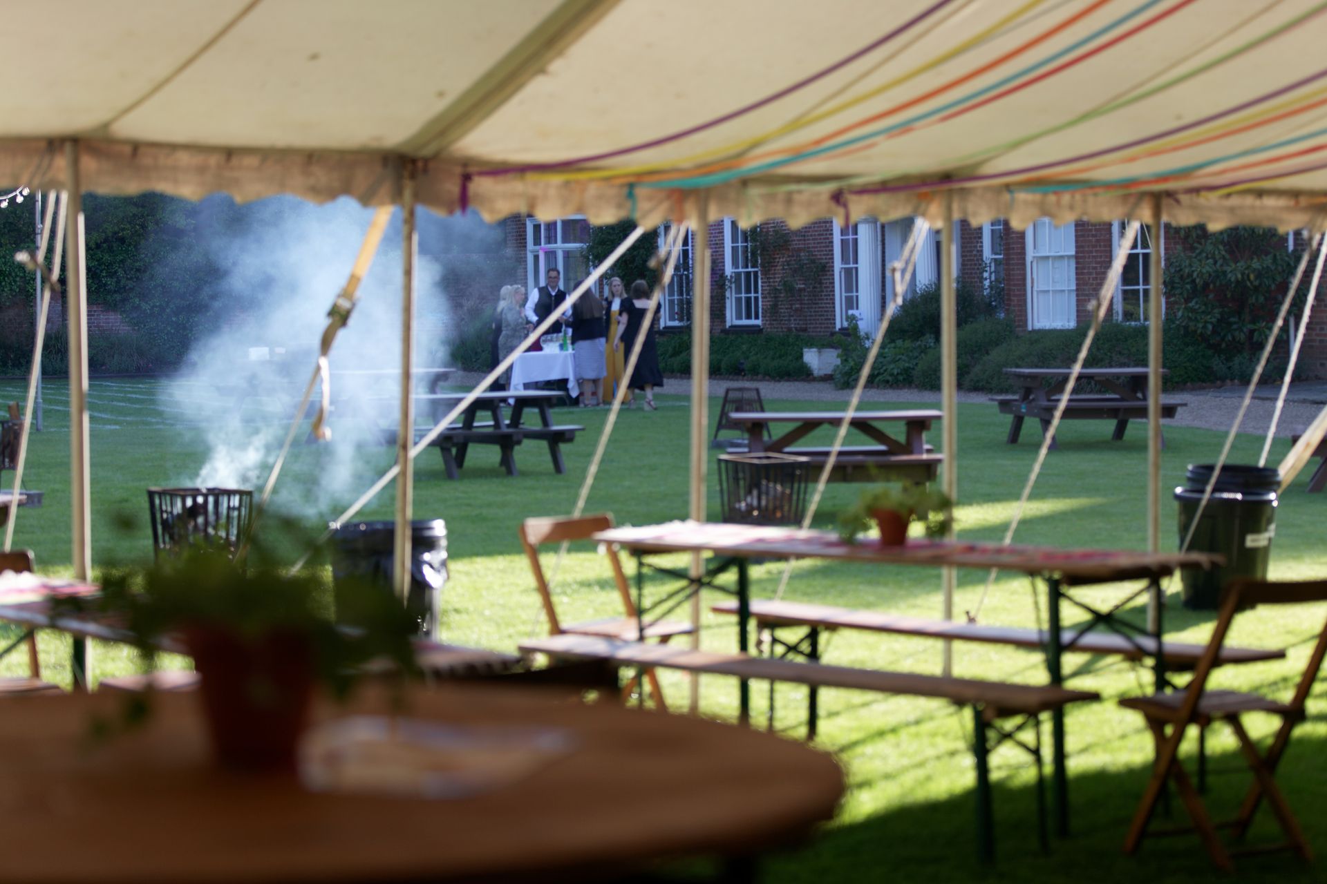 Outdoor event with tables and benches under a striped tent; a fire pit is lit in the background.