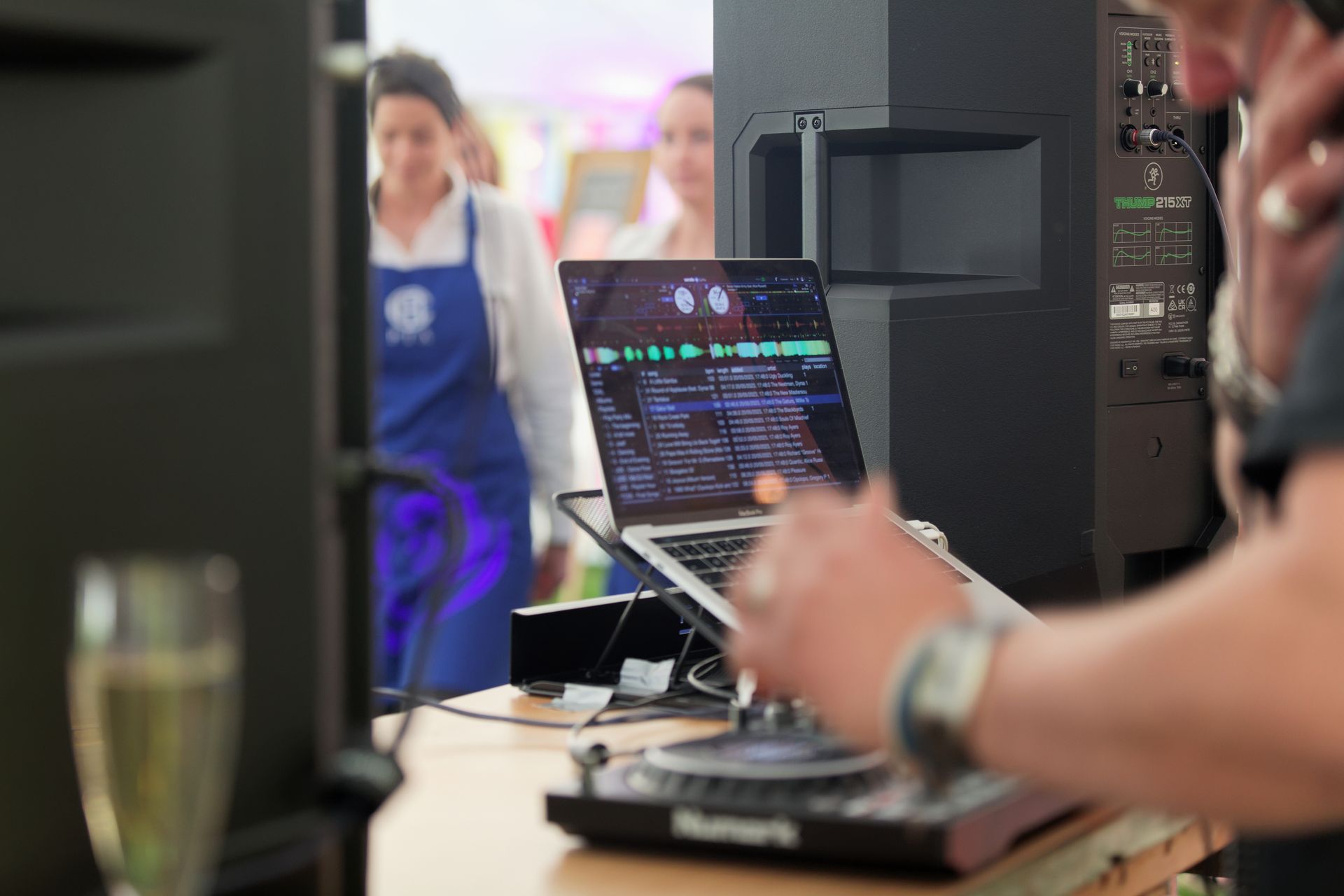 DJ at an event, mixing music on a laptop and controller. Two women in blue aprons in background.