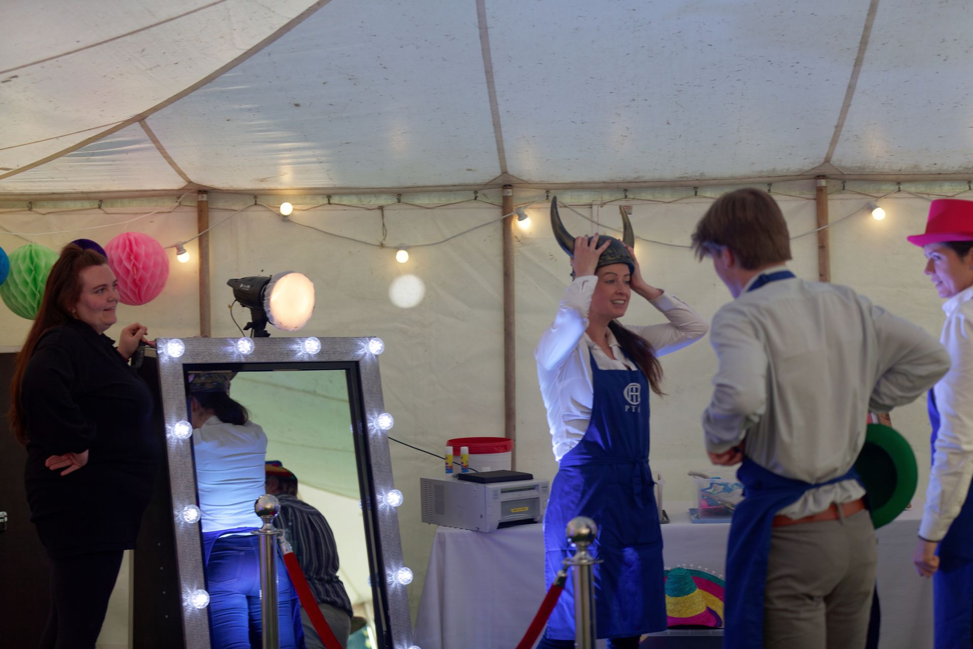 People at photo booth, woman wearing horns. Inside tent, fairy lights, hats.